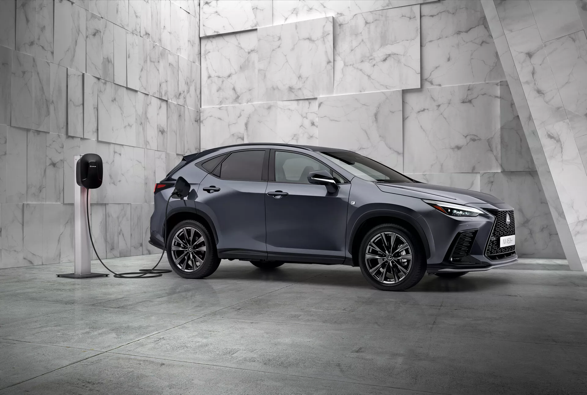 Esta imagen presenta el Lexus NX 2021, un SUV compacto moderno con un diseño elegante y deportivo, fotografiado desde una vista frontal de tres cuartos. Son visibles el lado izquierdo del vehículo y parcialmente su parte frontal, posicionados en diagonal dentro de un entorno minimalista adornado con grandes baldosas de mármol de color claro, sugiriendo una atmósfera premium. El Lexus NX presume de distintivos faros LED angulares y una parrilla frontal prominente, enfatizando su naturaleza sofisticada. Se observan retrovisores laterales aerodinámicos, llantas de aleación intrincadas de múltiples radios y manijas de puertas al ras que destacan su diseño deportivo y avanzado. Un detalle crucial es la conexión del automóvil a una estación de carga eléctrica, indicando que es un vehículo eléctrico o híbrido enchufable. El guardabarros delantero incluye un puerto de carga, indicando sus capacidades eléctricas. Esta imagen estática se centra en mostrar el diseño del vehículo y sus características tecnológicas en una composición equilibrada y tranquila con amplio espacio negativo, ejemplificando exclusividad e innovación en el diseño automotriz.