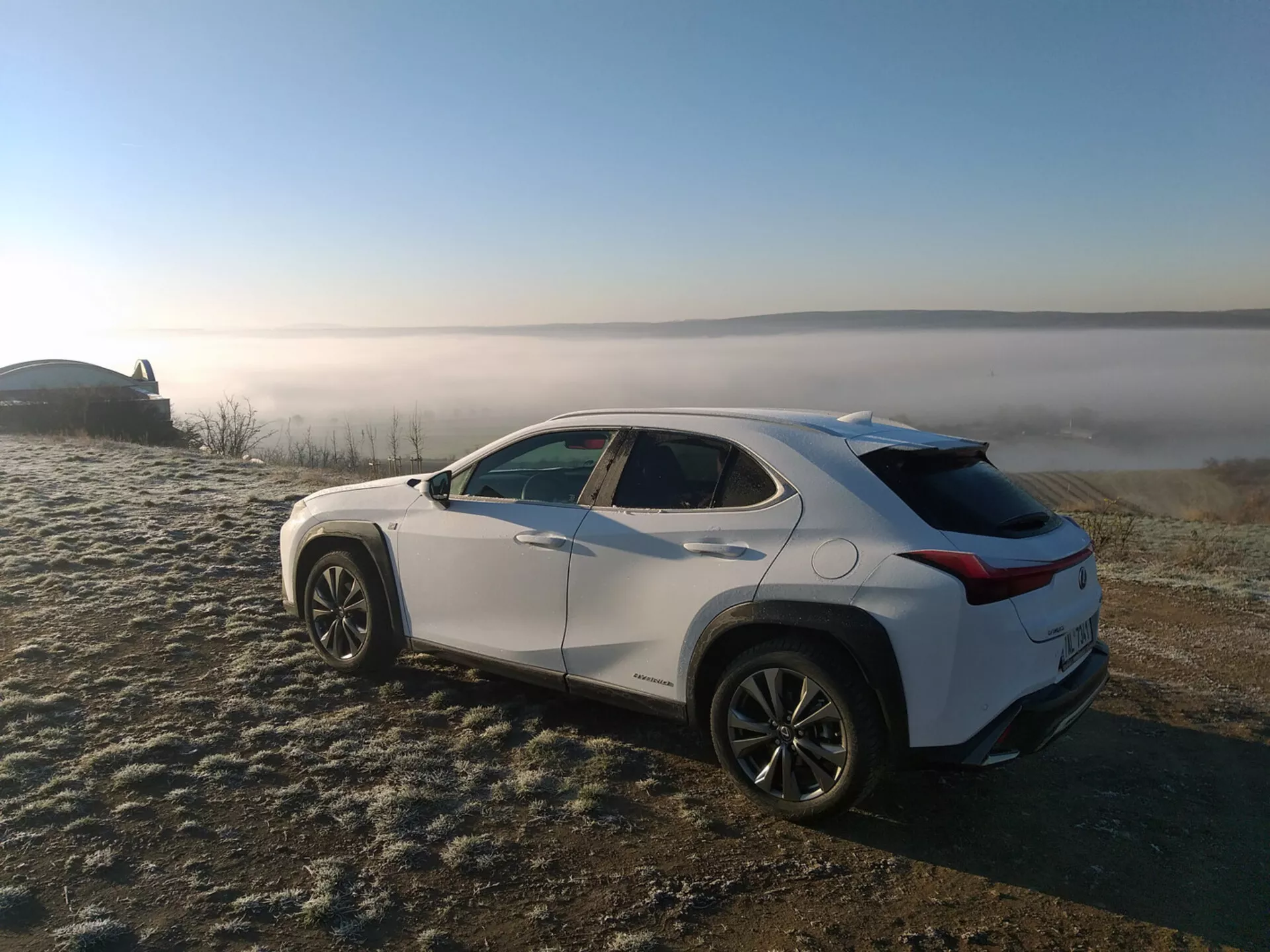 SUV crossover Lexus UX 2019 aparcado en hierba cubierta de escarcha con fondo de paisaje neblinoso, mostrando el perfil del vehículo desde un ángulo trasero de tres cuartos destacando las líneas de carácter lateral, arcos de rueda, línea de techo que se afina y llantas de aleación.