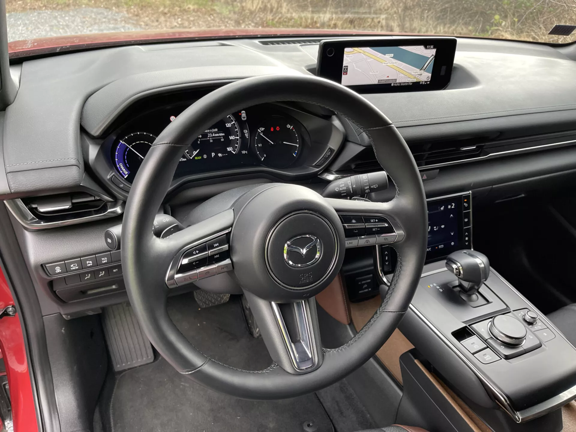 Vista interior del Mazda MX-30 2020 desde la perspectiva del conductor, enfocándose en detalles como el volante de tres radios envuelto en cuero con botones de control y levas de cambio, cuadro de instrumentos digital en el tablero con acentos azules, sistema de infotenimiento con pantalla táctil y navegación en la consola central, palanca de cambios deportiva, selector de modo de conducción, perilla de control adicional para el sistema de infotenimiento, lujosa mezcla de materiales con adornos negros y rojos, portavasos y paneles de control en la puerta para espejos laterales y ventanas, destacando la tecnología moderna y el diseño ergonómico.