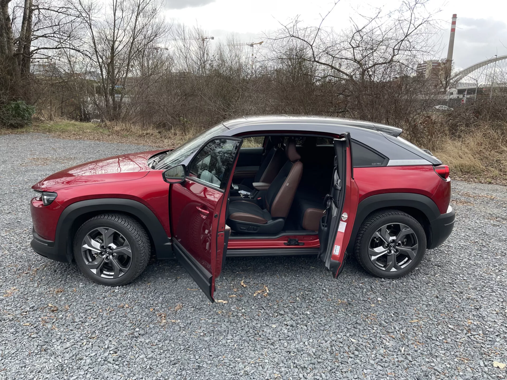 SUV Mazda MX-30 rojo de cuatro puertas de 2020 en perfil lateral sobre grava con puertas abiertas, ruedas de aleación de múltiples radios, ventanas tintadas, y techo de color contrastante contra un fondo industrial y de vegetación.