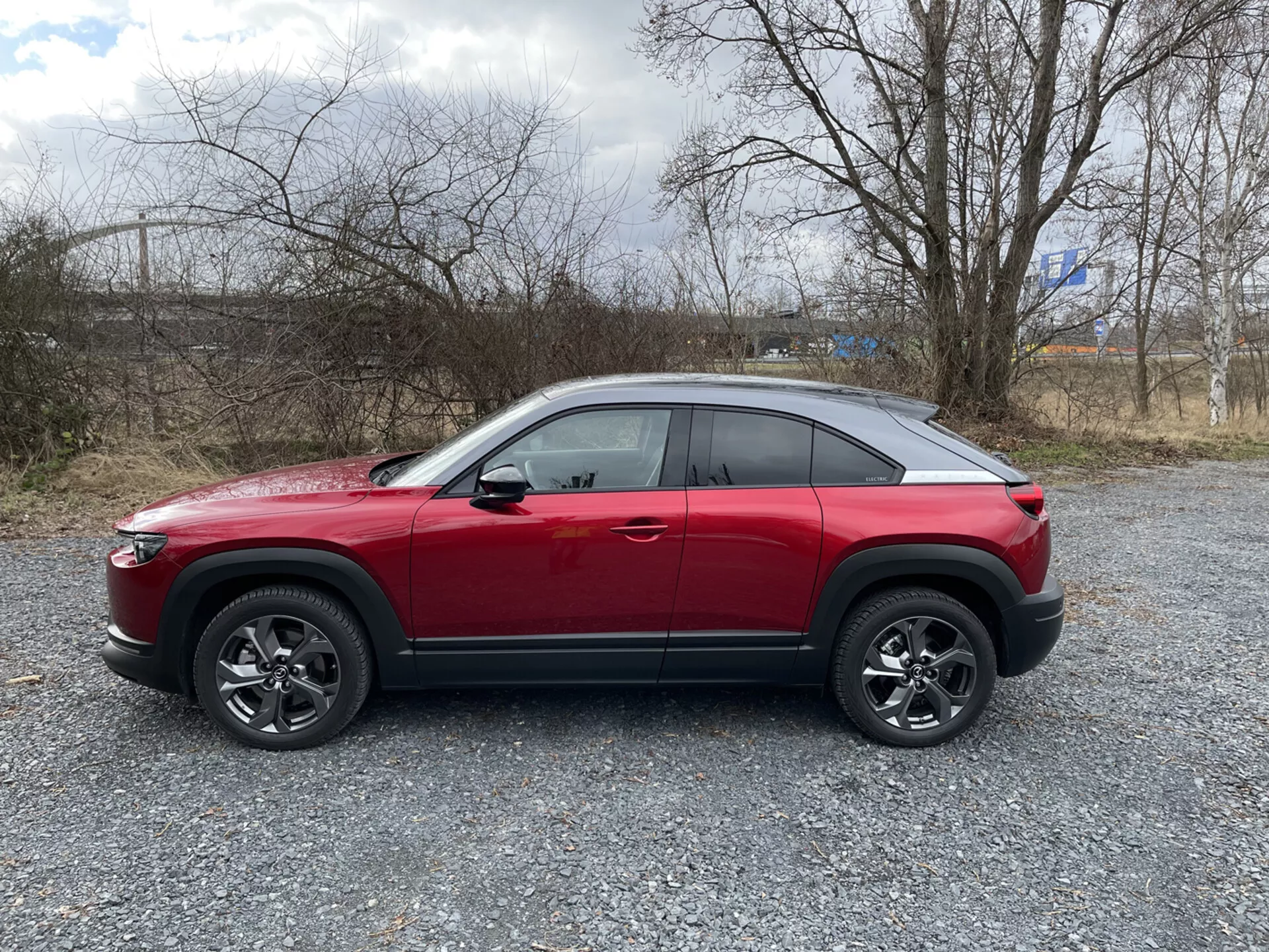 Perfil lateral del Mazda MX-30 rojo SUV crossover 2020 sobre grava con vegetación esparcida y cielo nublado, mostrando línea de cintura alta, línea de techo inclinada tipo cupé, línea de carácter dinámico, ruedas de aleación de multipalos con revestimiento negro, y posibles características tecnológicas avanzadas como ADAS, iluminación LED, entrada sin llave, TPMS y sistema de infotenimiento.