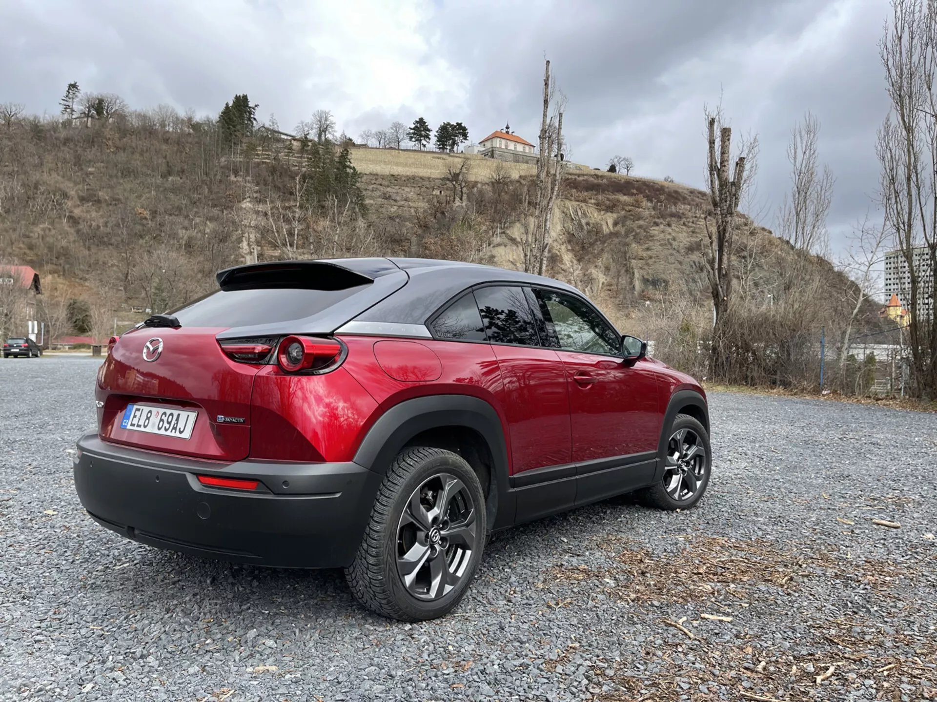 Crossover moderno Mazda MX-30 2020 estacionado en grava, mostrando su diseño deportivo en dos tonos rojo y negro, luces traseras LED, alerón trasero y ruedas de aleación de múltiples radios contra un fondo de naturaleza y arquitectura