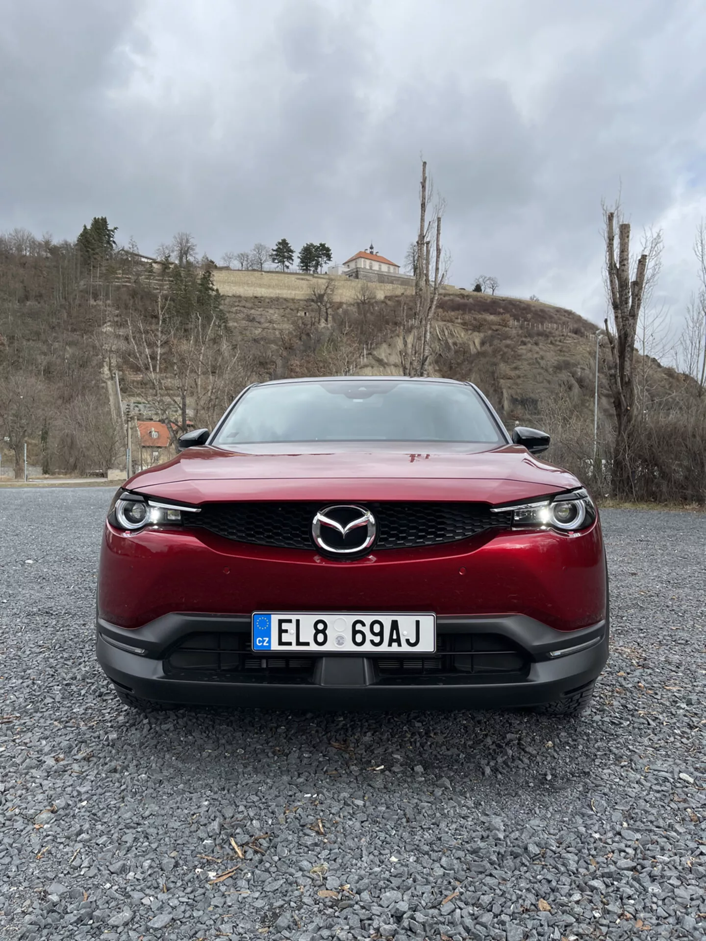 Vista frontal de un Mazda MX-30 de 2020 en rojo, destacando la parrilla del coche, el parachoques y los faros LED sobre una superficie de grava contra un fondo de cielo nublado con vegetación y un edificio