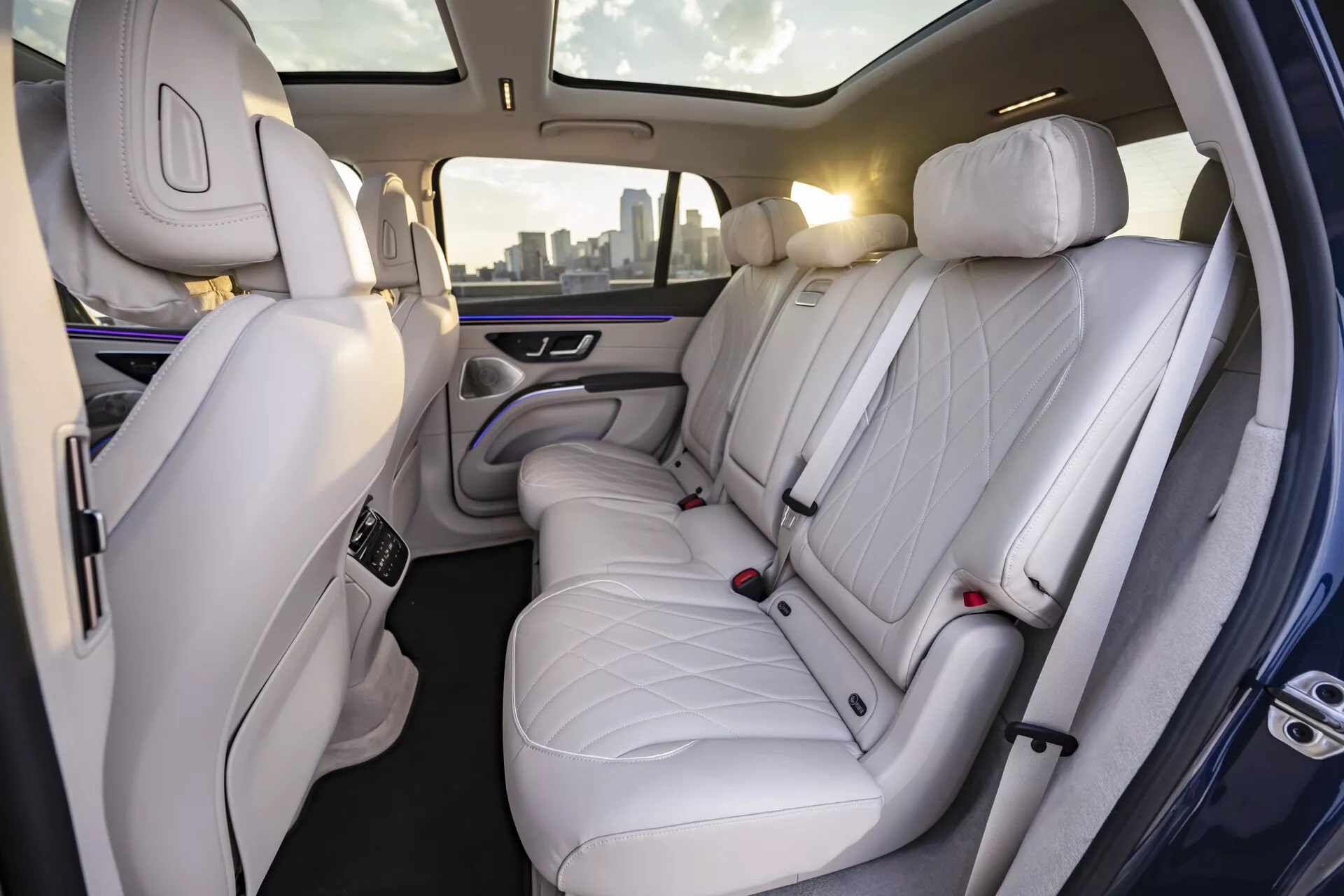 Vista del interior del Mercedes EQS-SUV 2022 que muestra el lujoso compartimento trasero para pasajeros. La imagen, tomada desde un ángulo ligeramente inclinado hacia el lado derecho del coche, revela asientos de cuero blanco o gris claro de alta calidad con un diseño de patrón de diamante acolchado. Con tres reposacabezas, los asientos brindan un amplio espacio, lo que indica un enfoque en la comodidad y la elegancia. También se puede ver la parte trasera del asiento del pasajero delantero, también tapizado en cuero premium. La sofisticada iluminación ambiental con un suave brillo púrpura a lo largo de los paneles de las puertas y el reposabrazos central mejora la sensación premium. El reposabrazos central cuenta con unidades de control, probablemente para ajustes climáticos y de asientos, y contiene dos portavasos.