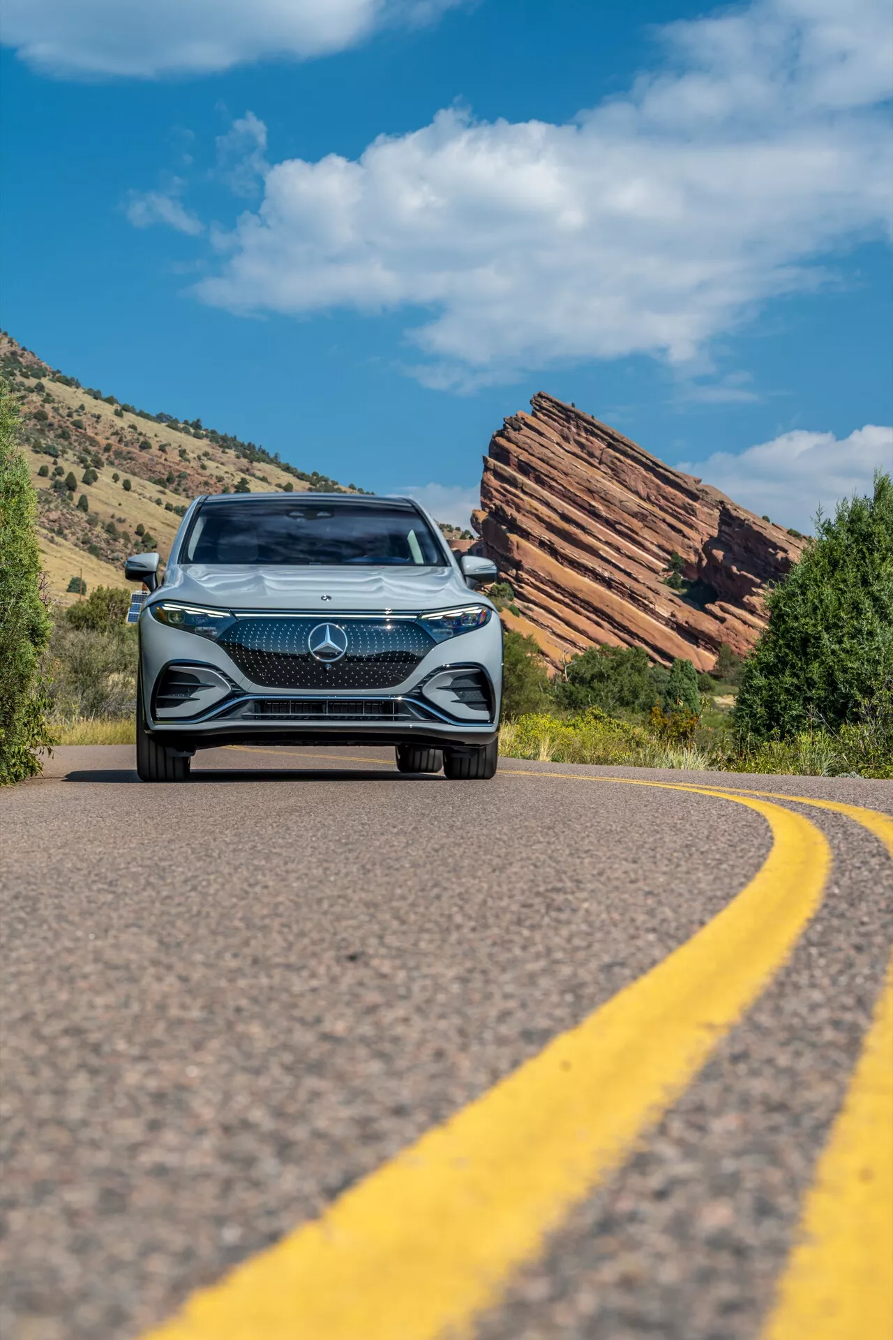 Toma en ángulo bajo de un Mercedes EQS SUV 2022 acercándose en una carretera curva, mostrando la audaz parrilla frontal con un gran emblema de estrella, diseño intrincado de diamantes, faros LED y luces diurnas, con un cielo azul claro y un terreno natural escarpado con formaciones rocosas estratificadas.