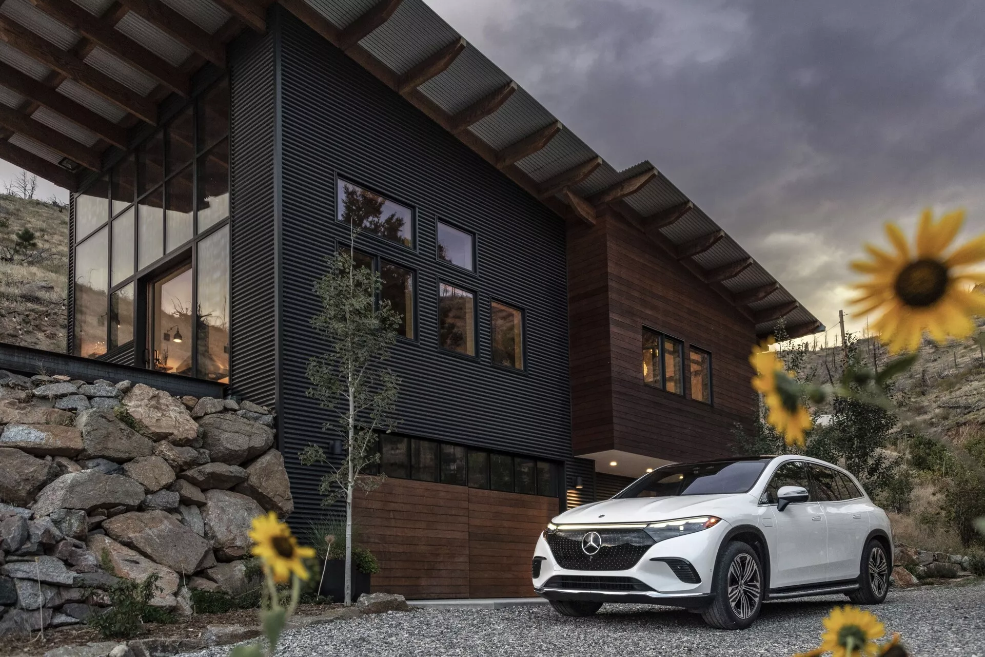 Imagen de un lujoso SUV Mercedes-Benz EQS 2022 estacionado frente a una casa contemporánea con un cielo nublado de fondo. La toma en ángulo bajo enfatiza la postura y el tamaño del vehículo, enfocándose en la parrilla delantera, el conjunto de faros, el parachoques delantero, los contornos del capó y el lado del conductor extendiéndose hasta la puerta trasera. Las características incluyen luces diurnas LED y ruedas elegantes, que añaden un sentido de lujo y tecnología avanzada. El SUV blanco contrasta con la casa oscura y el paisaje rocoso, rodeado de flores ligeramente desenfocadas debido a una poca profundidad de campo, destacando el SUV como el punto focal principal.