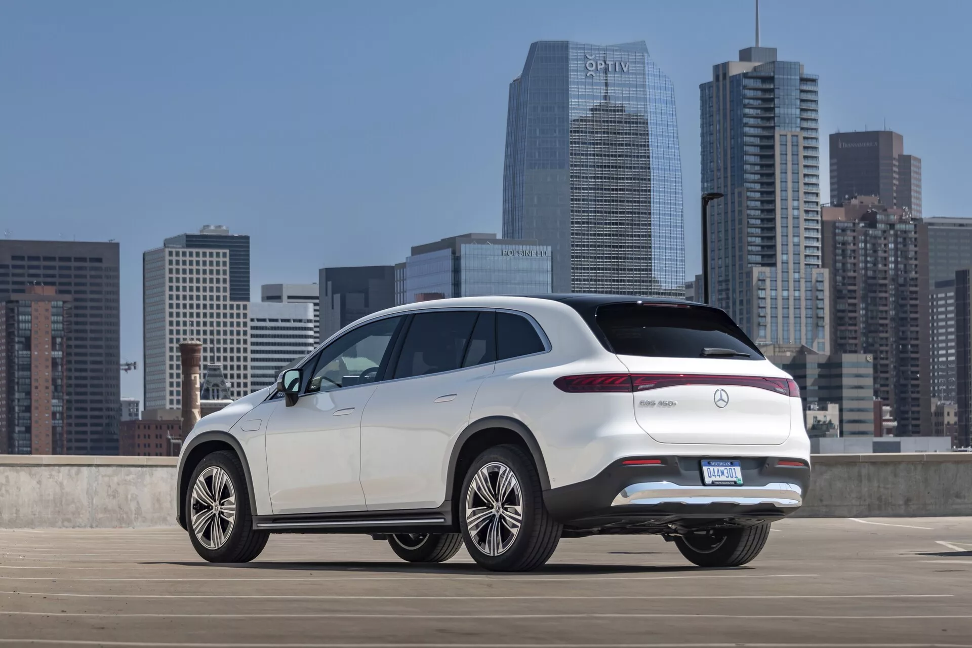 Vista trasera de tres cuartos de un Mercedes EQS-SUV 2022 blanco desde un ángulo ligeramente elevado, que muestra la parte superior de la sección trasera, el perfil lateral y características de diseño distintivas como luces traseras LED elegantes, portón trasero, parachoques trasero y grandes ruedas de aleación multirradios. El SUV está estacionado en una estructura de estacionamiento al aire libre con un horizonte urbano en el fondo, destacando su diseño moderno y de alta gama con posibles características avanzadas típicas de los modelos de lujo.