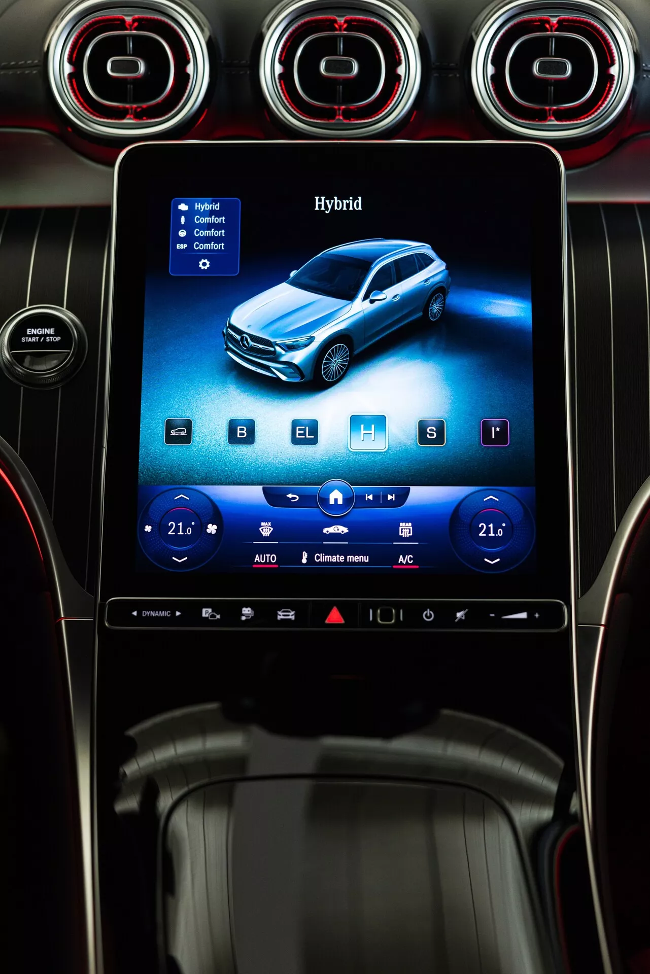 Vista de cerca de la consola central del Mercedes GLC 254 de 2022, con un sistema de infoentretenimiento con pantalla táctil vertical enmarcado por tres salidas de aire circulares con anillos de acento rojo. La pantalla muestra un vehículo híbrido azul, con controles de clima digitales ajustados a 21 grados Celsius en ambos lados, indicando un sistema de control de clima dual. Son visibles íconos para calefacción de asientos, enfriamiento y opciones de descongelación, junto con una barra de acceso rápido en la parte inferior. La consola está diseñada con acentos de metal cepillado, superficies negras brillantes y botones minimalistas, destacando un interior moderno y lujoso.