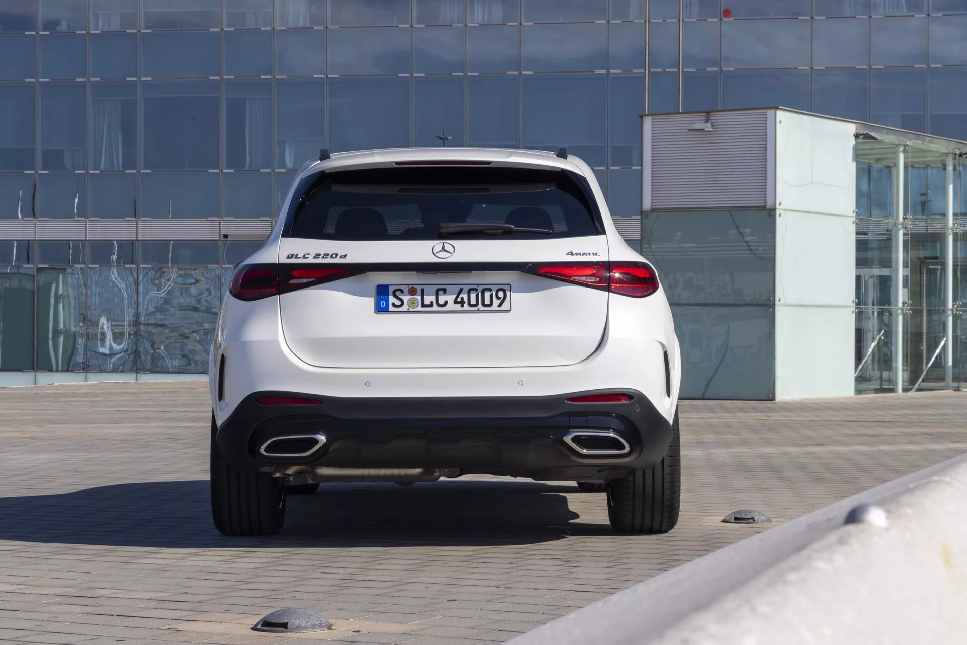 Vista trasera de un SUV compacto de lujo Mercedes-Benz GLC 300 del 2022 con capacidad de tracción en las cuatro ruedas 4MATIC, estacionado en un área pavimentada. La fotografía muestra la puerta trasera aerodinámica, el alerón trasero integrado, las luces traseras LED con un diseño horizontal, los dobles tubos de escape, los sensores de estacionamiento integrados, el difusor trasero y el acabado de alto brillo. El coche está centrado en el marco contra un fondo que presenta un edificio con fachada de vidrio, lo que indica un entorno urbano o comercial. La imagen bien iluminada destaca las características de diseño elegante y deportivo típicas de los vehículos modernos de Mercedes-Benz.