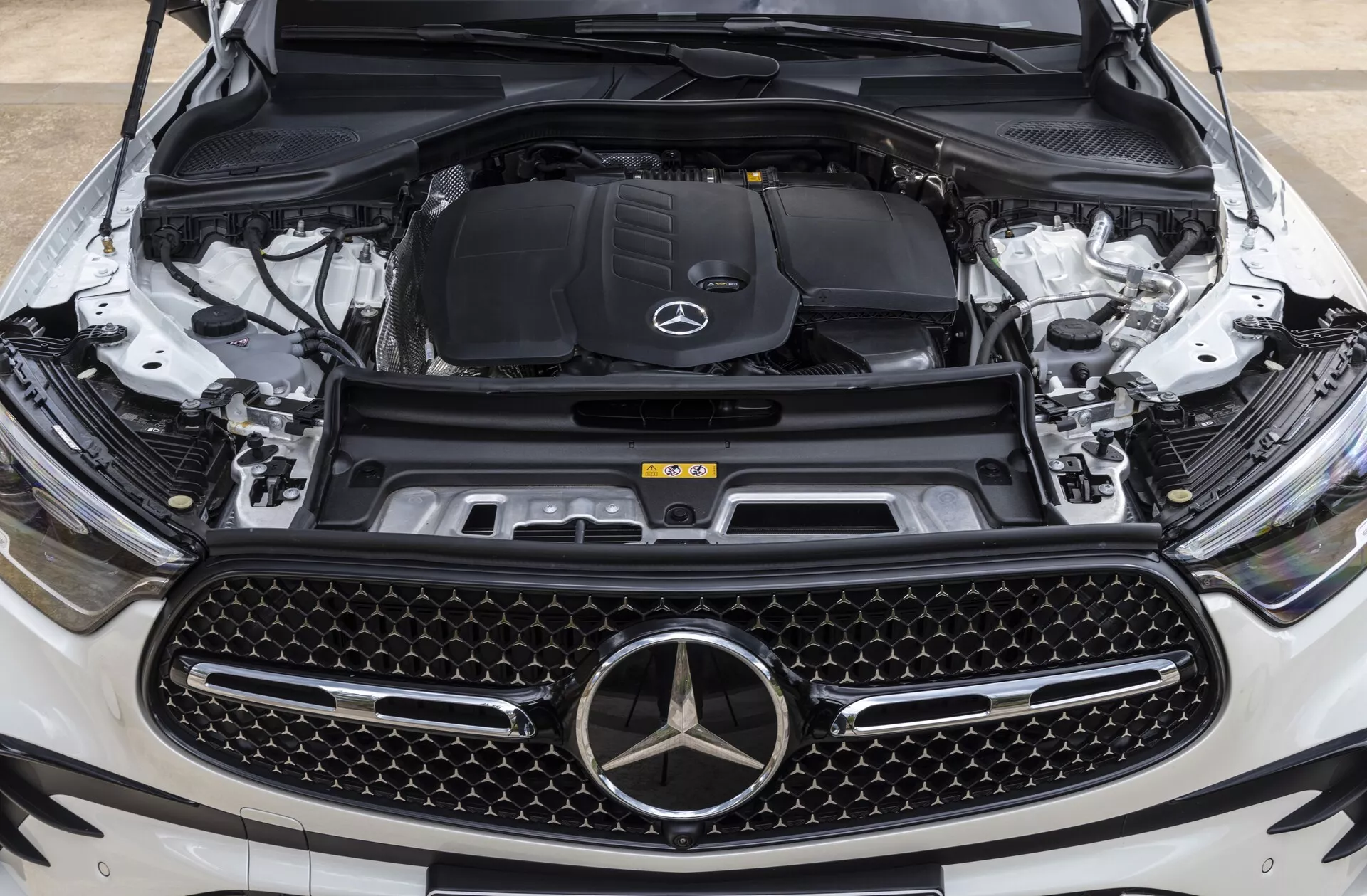 Vista frontal en primer plano del Mercedes-Benz GLC 2022 (254) con el capó abierto mostrando el compartimento del motor desde una perspectiva al nivel de los ojos centrada en el emblema de la marca y la parrilla, destacando la gran parrilla con patrón de diamante, el emblema de estrella de Mercedes-Benz, el compartimento del motor con el logotipo y componentes visibles como el bloque del motor, el sistema de admisión de aire, el depósito de refrigerante, la torre de suspensión y la cámara frontal, indicando características tecnológicas avanzadas.