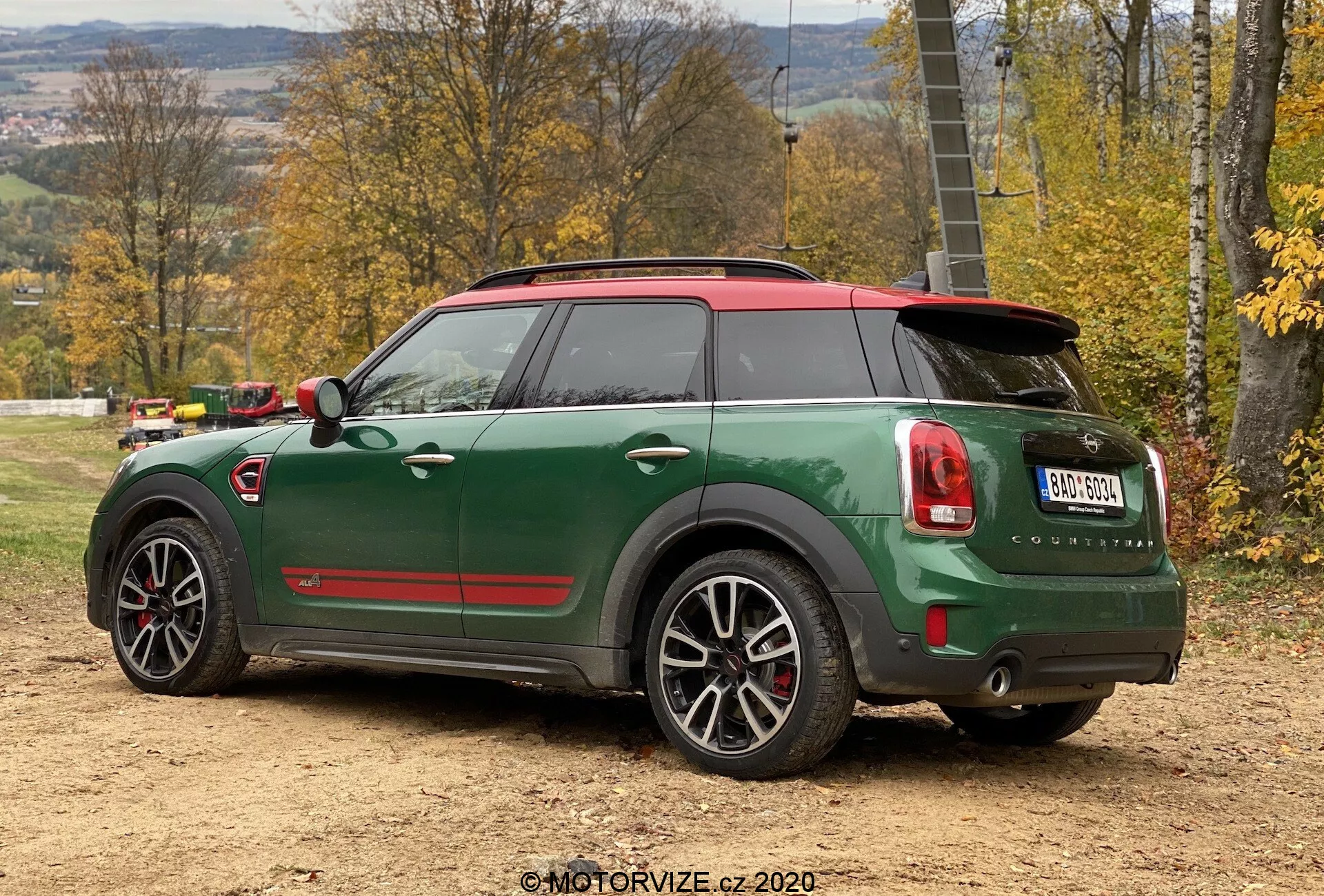 Vista trasera de tres cuartos del Mini Cooper Countryman verde 2020 con edición deportiva John Cooper Works, mostrando techo rojo y tapas de espejo, doble salida de escape, alerón trasero, llantas de aleación de cinco radios con detalles en negro y características tecnológicas avanzadas como sensores de estacionamiento y antena de aleta de tiburón, colocado en una superficie de tierra en un entorno rural otoñal.
