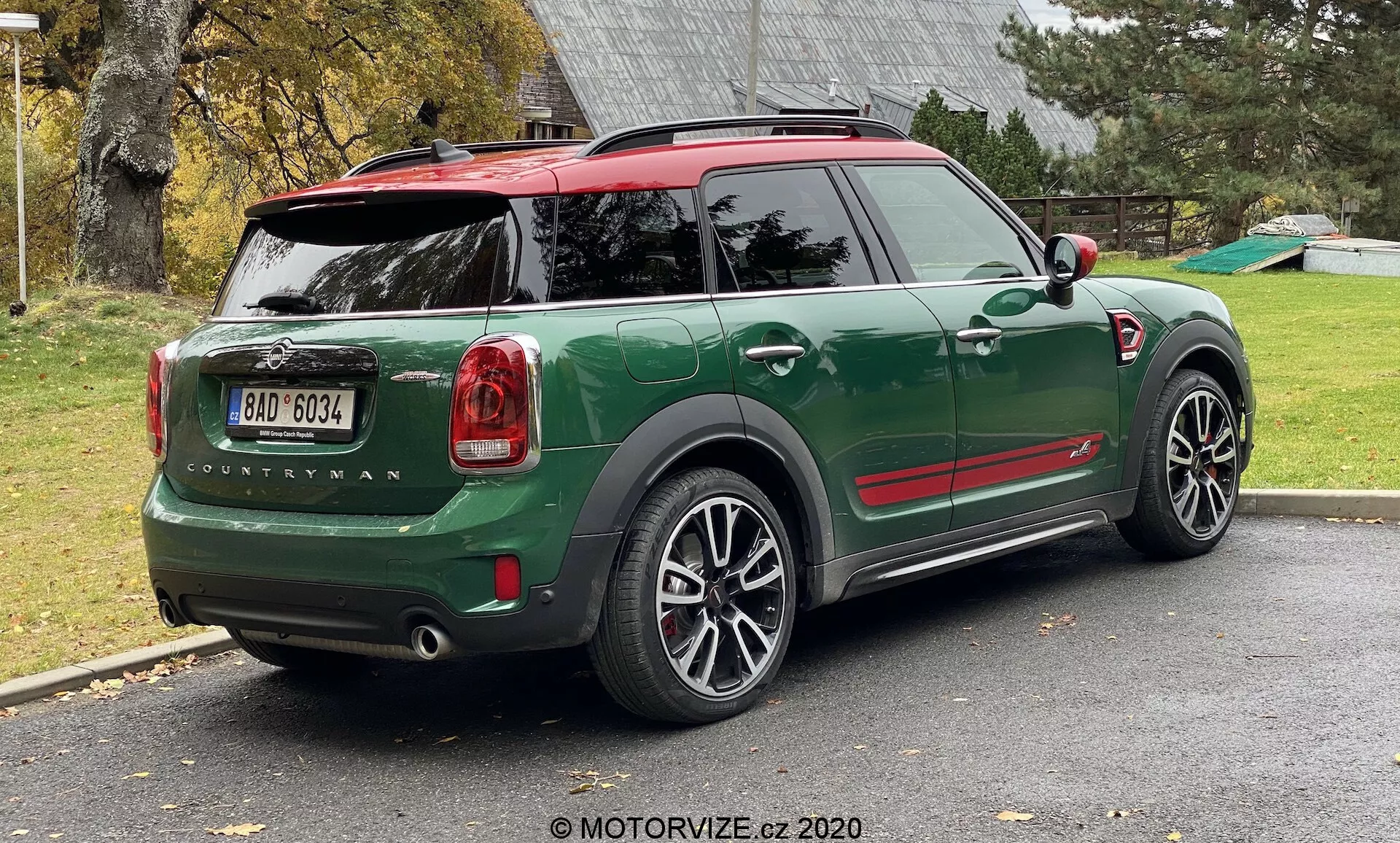 Vista trasera de tres cuartos de un MINI Countryman John Cooper Works 2020 verde estacionado en una entrada residencial. La imagen muestra la mitad trasera completa del lado derecho del coche, incluidas la puerta trasera, la rueda trasera derecha, parte del techo y las puertas de los pasajeros. El diseño deportivo y compacto del SUV presenta acentos rojos contrastantes en las carcasas de los espejos laterales y el techo, arcos de rueda negros y grandes ruedas de aleación de varios radios. Las luces traseras, el nombre del modelo 'Countryman' y el logotipo de MINI son claramente visibles. El coche tiene un techo rojo con un alerón y raíles en el techo, y la ventana trasera incluye un descongelador y un limpiaparabrisas.
