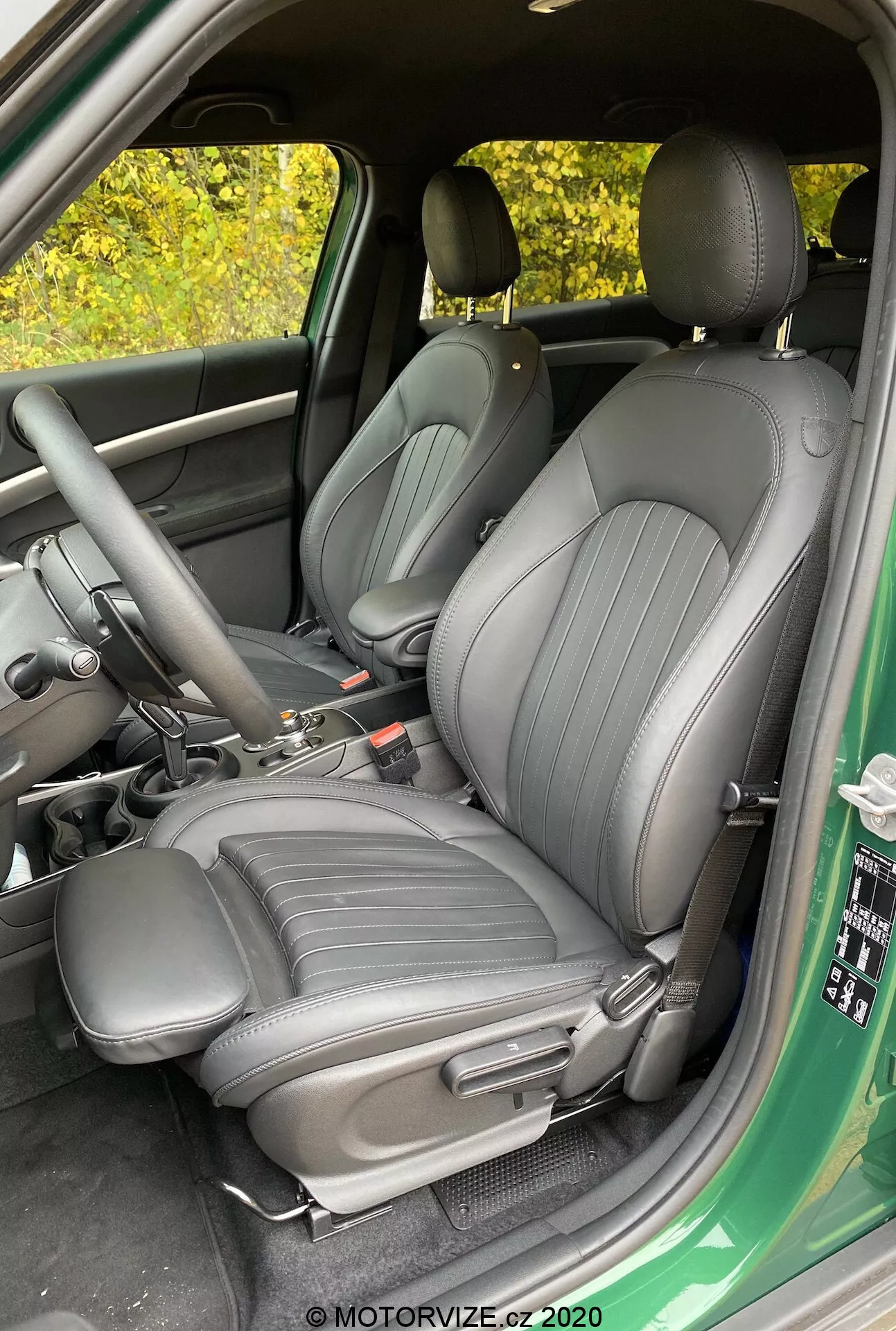 Imagen del interior del Mini Countryman John Cooper Works 2020 centrada en los asientos delanteros del conductor y el pasajero, tapizados en un material oscuro de alta calidad con líneas verticales, vista desde la puerta del conductor abierta, mostrando la consola central con la palanca de cambios y el freno de mano, vista parcial del panel de la puerta con controles para las ventanas y espejos.
