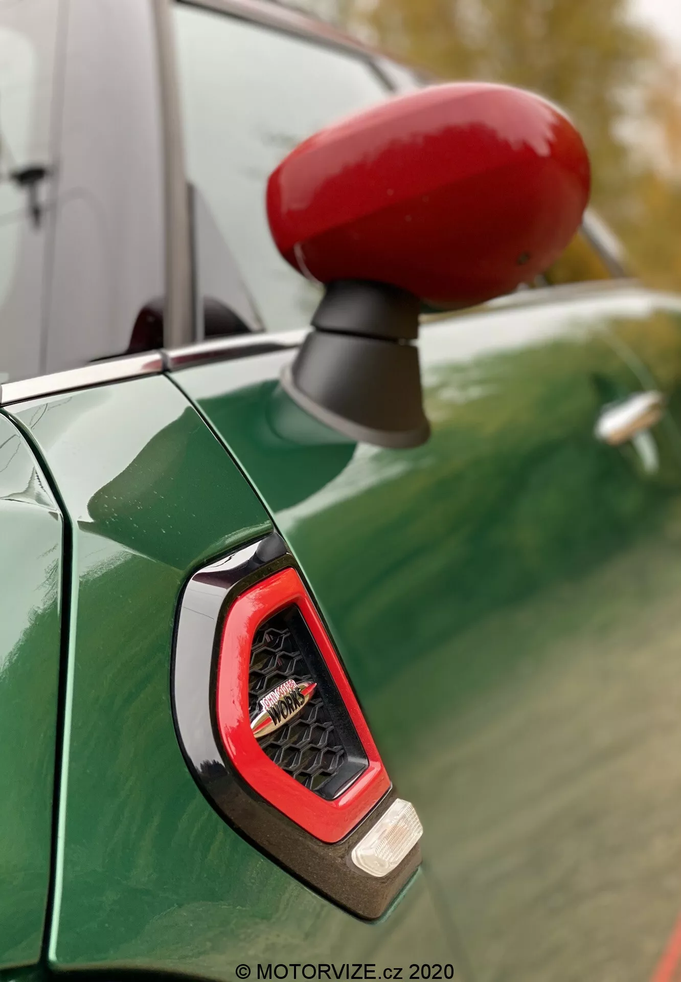 Primer plano del espejo lateral de color rojo vibrante y el indicador lateral del lado del conductor de un Mini Countryman John Cooper Works 2020, visto contra un panel de carrocería verde metálico profundo con reflejos de un cielo nublado y árboles. El enfoque resalta el indicador angular con lente de cristal probablemente mostrando tecnología LED, rodeado por un acabado deportivo y brillante.