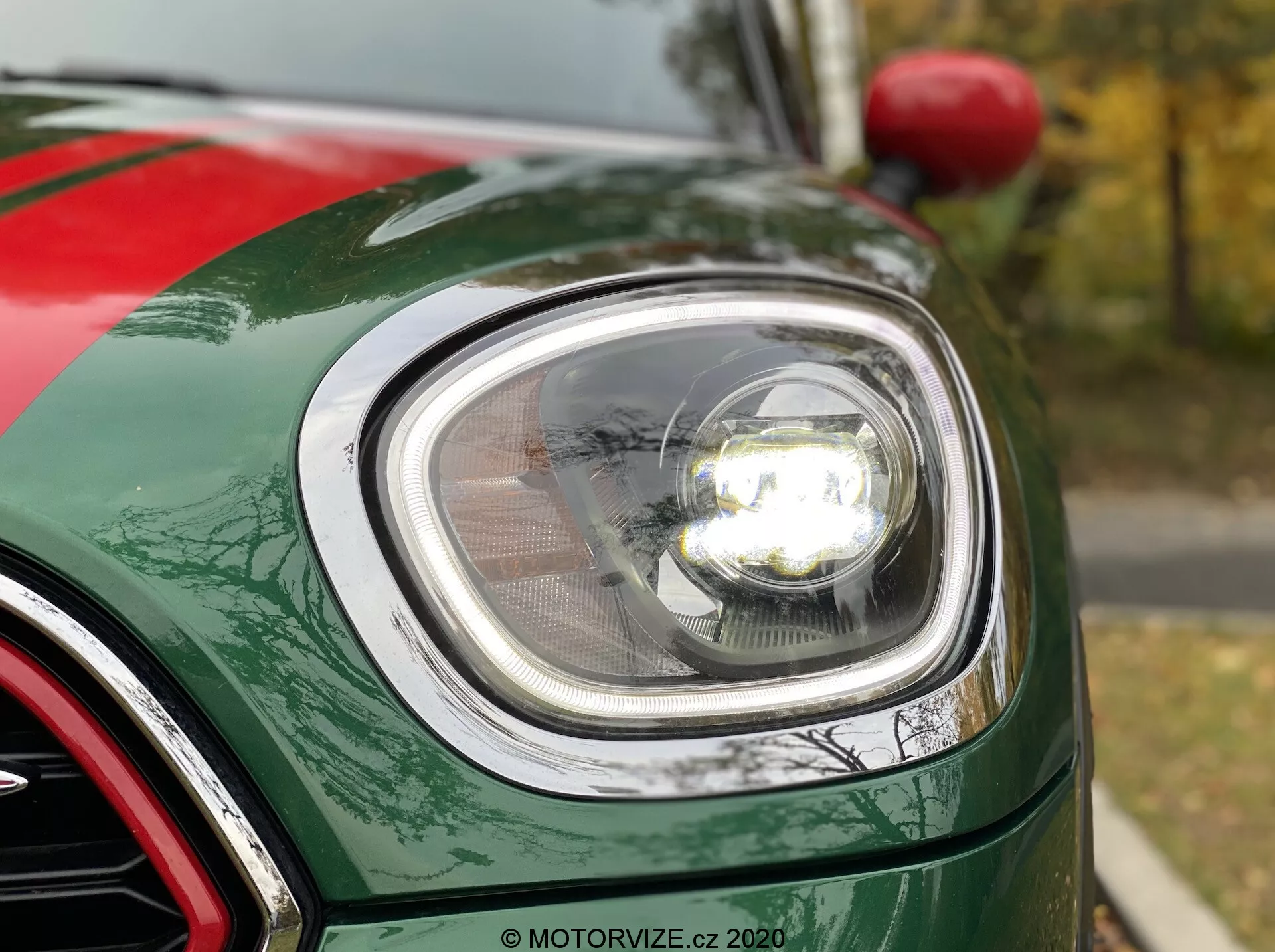 Vista de cerca de la sección frontal izquierda de un Mini Countryman John Cooper Works 2020, enfocándose en el moderno faro redondo con tecnología LED, cubierta de lente clara y faro estilo proyector iluminado. La imagen muestra la curvatura del guardabarros delantero, el borde del capó y parte de la parrilla acabada en cromo. El coche tiene un acabado de pintura verde metálica brillante con escamas metálicas visibles.