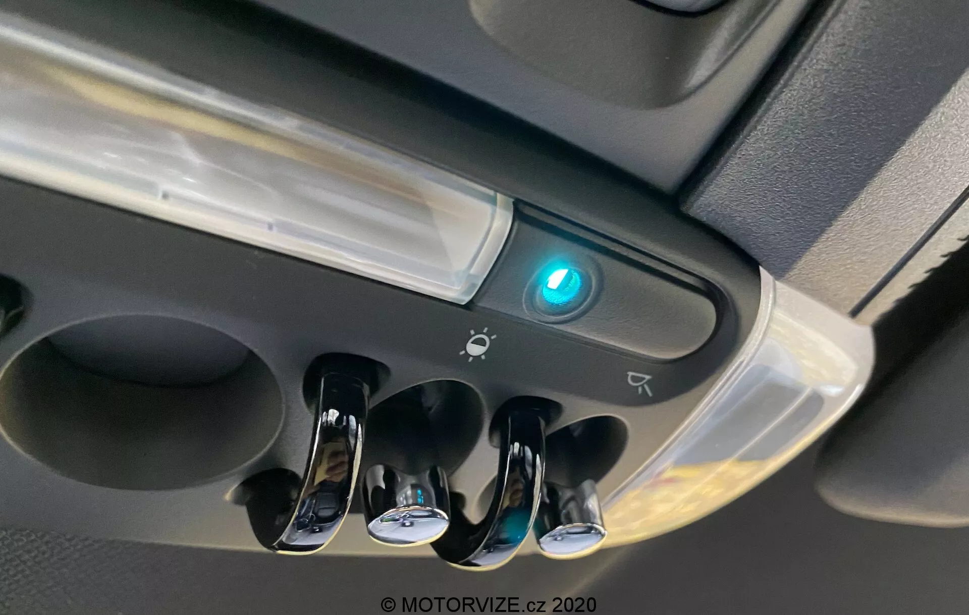 Vista de cerca del área de la consola superior del Mini Countryman 2020 John Cooper Works que muestra las luces interiores de la cabina, tres interruptores con acabado cromado para luces de lectura individuales, una luz central de cabina, un botón iluminado en azul que podría indicar conectividad Bluetooth o una característica de seguridad, el borde de un posible compartimento de almacenamiento, iconos que sugieren controles adicionales o sensores y un diseño moderno con superficies brillantes y mate.
