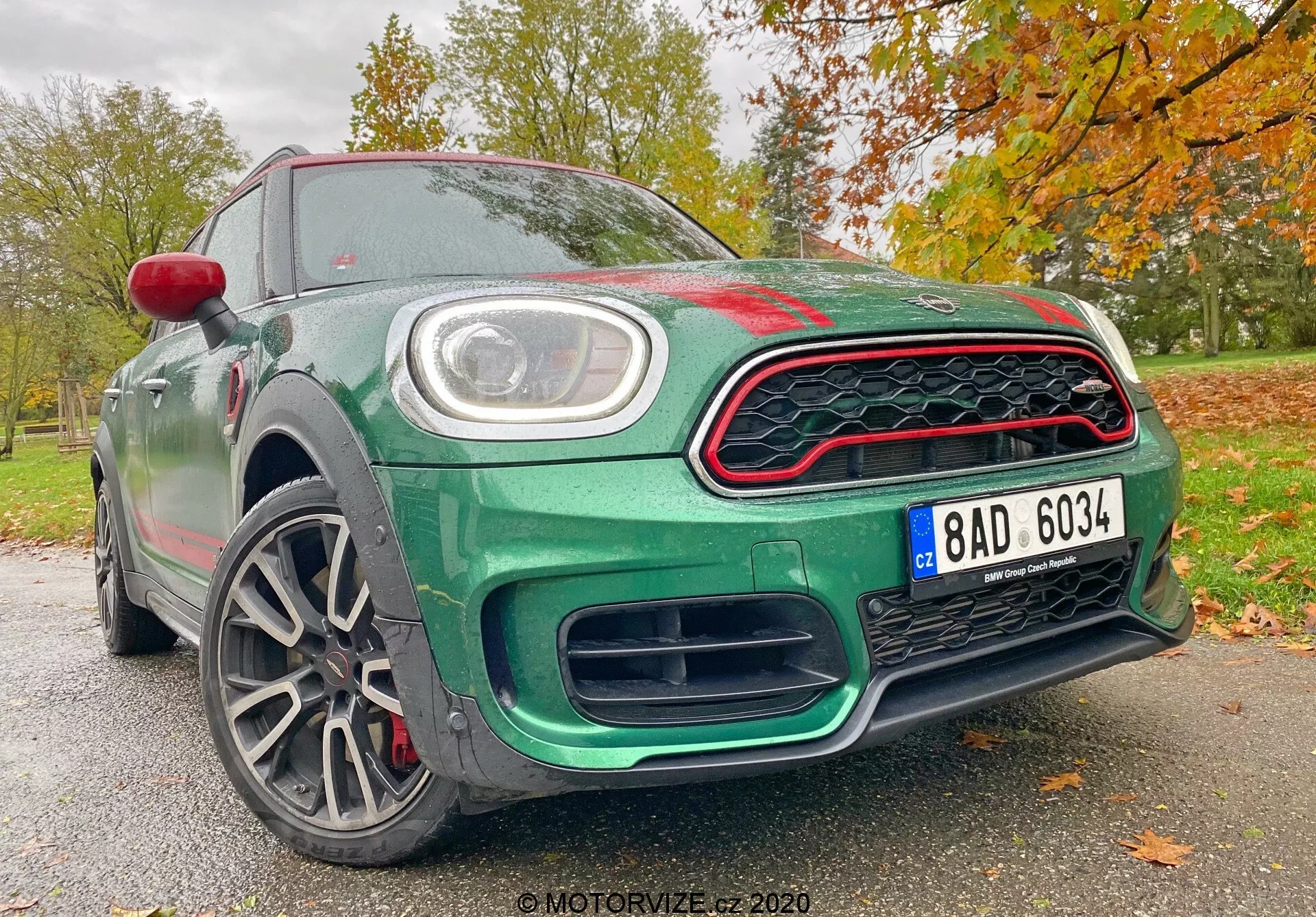 La imagen muestra el Mini Countryman 2020 edición John Cooper Works desde un ángulo frontal bajo, resaltando su diseño deportivo y moderno. Los elementos visibles incluyen la parrilla frontal de patrón de panal con acentos rojos, grandes entradas de aire debajo de la parrilla para mejorar el flujo de aire, una matrícula central y luces diurnas LED circulares incrustadas en los faros principales. El capó muestra una ligera protuberancia con pliegues aerodinámicos, mientras que las líneas esculpidas en el perfil lateral mejoran su apariencia dinámica. Las llantas de aleación de múltiples radios sugieren una construcción ligera para un mejor manejo y aceleración, y revelan pinzas y discos de freno de alto rendimiento. La carrocería está pintada en un rico verde metálico, acentuado por una tapa de espejo lateral roja y acentos rojos en la parrilla, contra un fondo otoñal de hojas. Esta perspectiva enfatiza las características tecnológicas y de diseño avanzado del automóvil, haciéndolo destacar en la categoría de vehículos deportivos y de lujo.