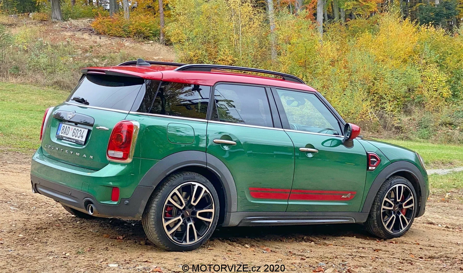 Vista trasera de tres cuartos de un Mini Countryman John Cooper Works 2020 parado en una superficie sin pavimentar con parches de césped. El compacto deportivo presenta un esquema de colores dual con una carrocería verde oscuro y un techo y espejos rojos contrastantes. El diseño trasero muestra una puerta trasera, limpiaparabrisas trasero, insignia prominente y luces traseras dispuestas verticalmente. El parachoques trasero incluye sensores de estacionamiento. También cuenta con ruedas de aleación deportivas con diseño de múltiples radios que muestran los frenos, sugiriendo una versión de alto rendimiento. El fondo natural con árboles otoñales insinúa la estación.