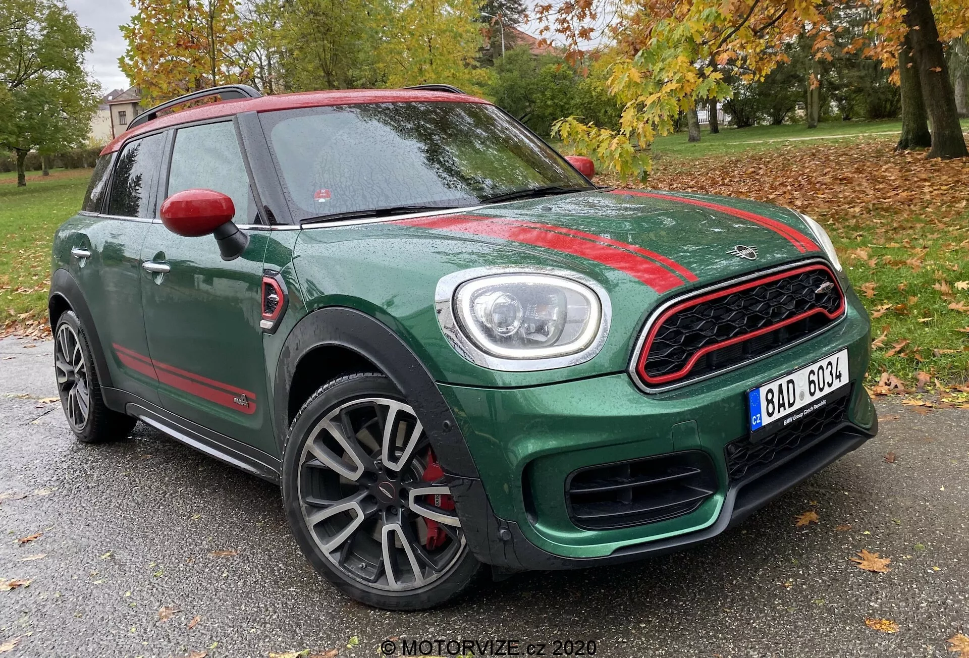 Vista de tres cuartos delantero de un Mini Countryman John Cooper Works 2020, mostrando su carrocería compacta SUV verde con acentos rojos contrastantes. Capturada desde un ángulo bajo, la imagen resalta la parrilla frontal deportiva con un borde cromado y diseño de panal, faros circulares prominentes con luces diurnas LED, llantas de aleación multirradio con acabado gris y tapas de retrovisores rojas. La foto está ambientada con un fondo de árboles y una estructura tenue, sugiriendo una ubicación en un parque o área residencial.