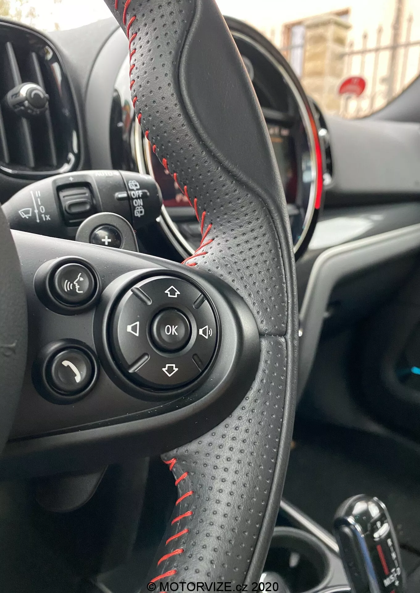 Vista interior de un Mini Countryman John Cooper Works 2020 centrada en el volante revestido de cuero con costuras rojas, botones de control para audio y llamadas, y un cuadro de instrumentos parcialmente visible con acentos rojos que indican un modelo deportivo. Muestra el área de control del conductor, la palanca de cambios automática y los controles de las ventanas eléctricas, destacando la conveniencia y las funciones avanzadas.