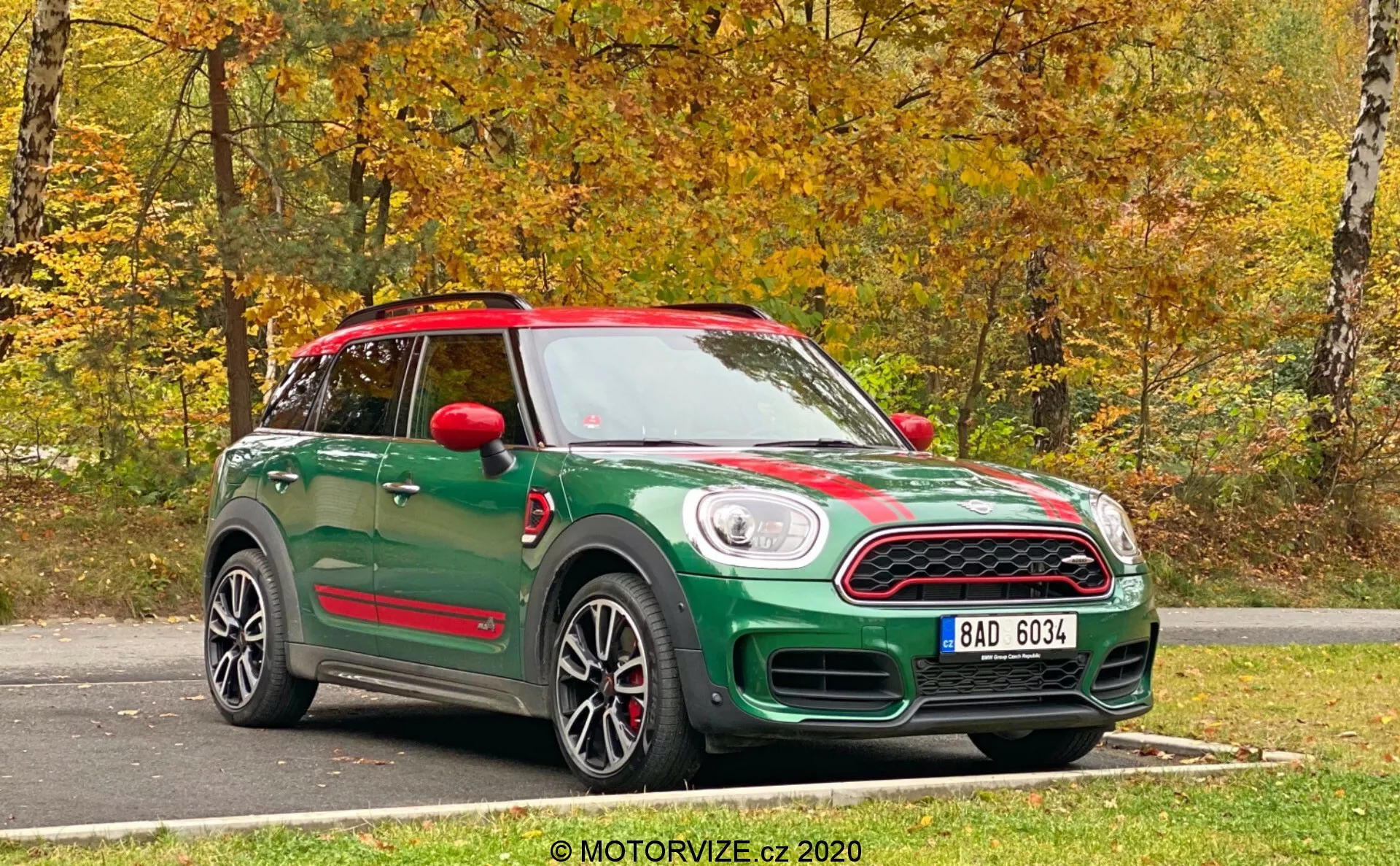 Un Mini Countryman John Cooper Works 2020 verde capturado desde una vista frontal de tres cuartos. La fotografía muestra tanto la parte delantera como el lateral del coche, destacando sus características de diseño. La parte delantera tiene una parrilla distintiva con una matrícula debajo y faros redondos clásicos. Rayas deportivas corren a lo largo del capó, y el perfil lateral muestra llantas de aleación, espejos laterales y la línea del techo. Los espejos laterales rojos y el techo a juego contribuyen a un esquema de color bicolor. Debajo de las puertas, los estribos laterales coordinados sugieren un paquete de nivel de acabado superior o deportivo. El vehículo tiene un techo solar panorámico y está estacionado en una superficie de asfalto con un fondo natural de otoño. La foto nítida y brillante fue tomada con luz natural.