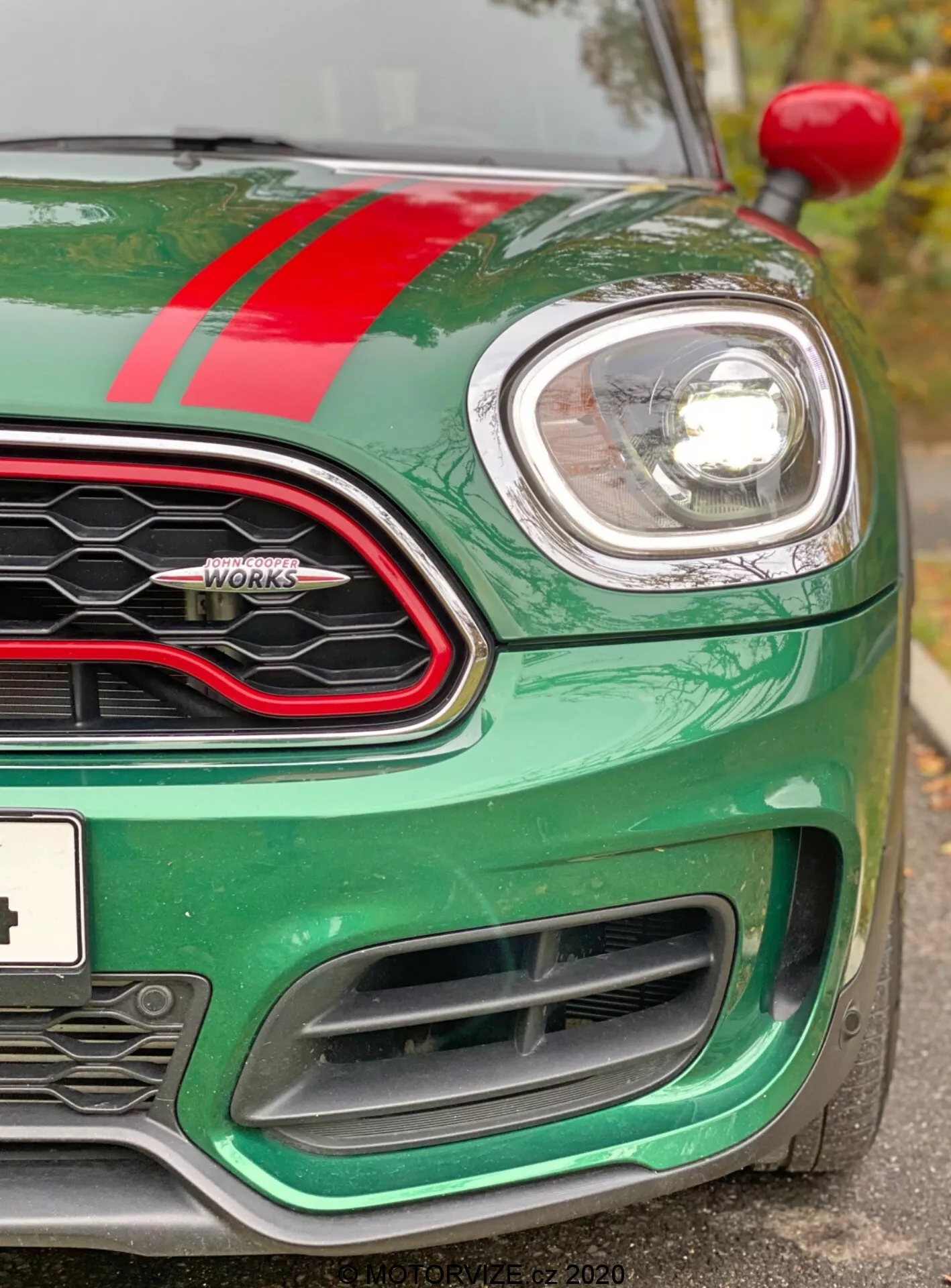 Vista detallada de la esquina delantera izquierda de un Mini Countryman John Cooper Works 2020. La imagen, tomada ligeramente por debajo del nivel de los faros, muestra el capó verde metálico con una franja de carreras roja, el faro izquierdo circular envuelto en cromo y una parte de la parrilla de panal con un borde negro brillante. Se puede ver la insignia de 'John Cooper Works', el indicador izquierdo en el arco de la rueda y las grandes entradas de aire del parachoques delantero diseñadas para enfriar el motor y mejorar la aerodinámica. El coche aparece limpio y bien mantenido, destacando su tecnología de faros modernos de haz de proyector o LED y su diseño deportivo y orientado al rendimiento.