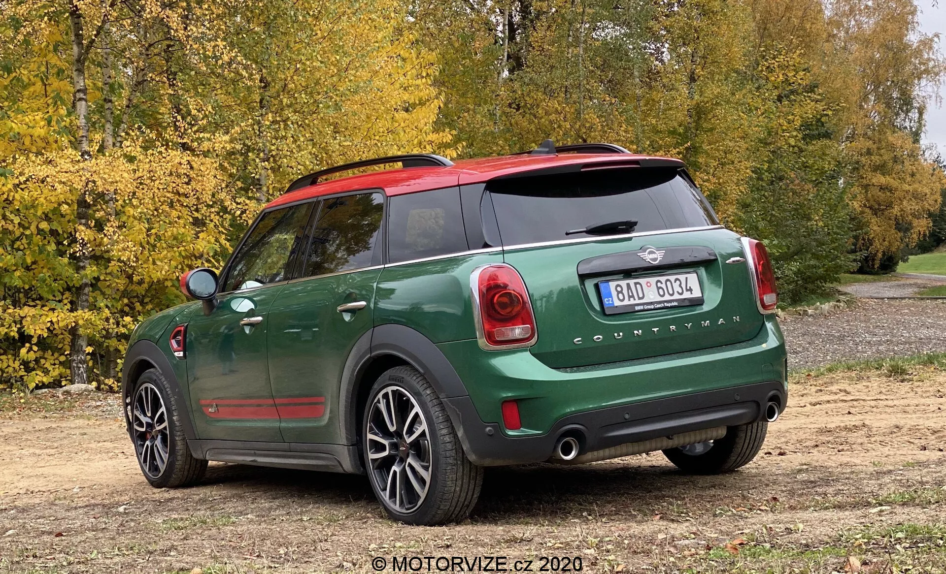 Vista trasera de tres cuartos de un Mini Cooper Countryman John Cooper Works 2020 con una pintura de dos tonos verde oscuro y negro, tapas de espejos rojas, emblema de Mini visible y letras 'Countryman', luces traseras de diseño único, parachoques trasero prominente con sensores de estacionamiento, ventana trasera con desempañador, tubos de escape dobles, limpiaparabrisas trasero, llantas de aleación de cinco radios, estacionado en un camino de tierra con árboles y hojas de otoño en el fondo, capturado desde un ángulo ligeramente alto.