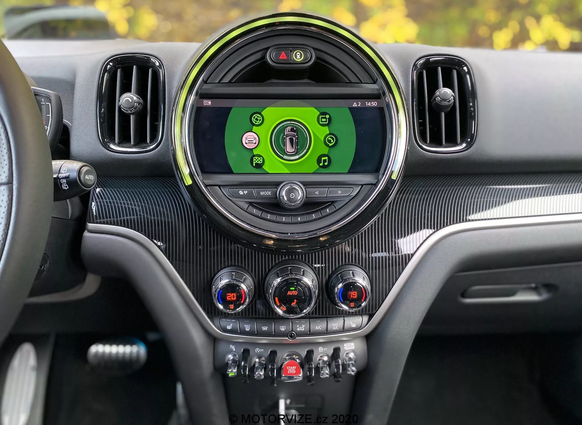 Panel de instrumentos interior del Mini Countryman John Cooper Works 2020 desde la perspectiva del conductor. Consola central con pantalla de infoentretenimiento circular grande mostrando iconos coloridos para medios, conectividad telefónica, navegación y configuraciones del vehículo. Enmarcado por un bisel circular elegante y flanqueado por ventilaciones adicionales. Debajo de la pantalla hay diales analógicos con acentos cromados para el control del clima y otros interruptores para funciones como calefacción de asientos, desempañado, recirculación de aire y modos de conducción. Textura tipo fibra de carbono y acentos metálicos proporcionan un diseño interior moderno y lujoso con clúster de instrumentos digital visible indicando que el vehículo está encendido.