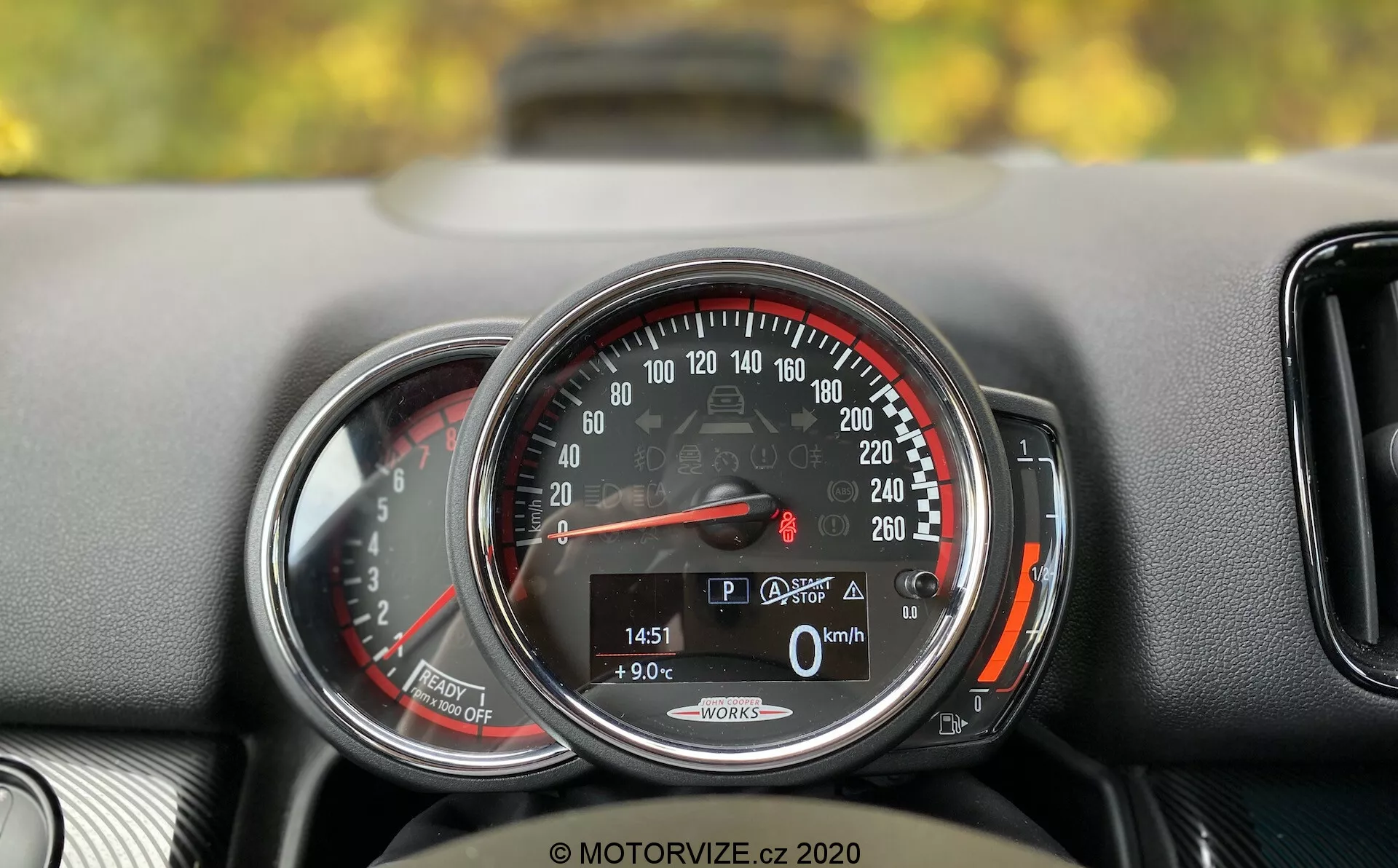 Primer plano detallado del panel de instrumentos del Mini Countryman John Cooper Works 2020. El enfoque está en un velocímetro circular que va de 0 a 260 km/h, con la aguja señalando 0 km/h, indicando que el coche está parado. Una pantalla digital en la parte inferior muestra el coche en modo 'P', una temperatura exterior de 9.0°C, un kilometraje de 1451 km y el icono de maletero abierto. Las palabras 'Auto Start/Stop' sugieren un sistema de ahorro de combustible. El velocímetro está enmarcado por un anillo cromado y tiene una zona roja comenzando justo después de 220 km/h. A la izquierda, hay una vista parcial de otro indicador, probablemente un tacómetro. El salpicadero tiene una superficie texturizada y una pequeña ventilación es parcialmente visible a la derecha del velocímetro.
