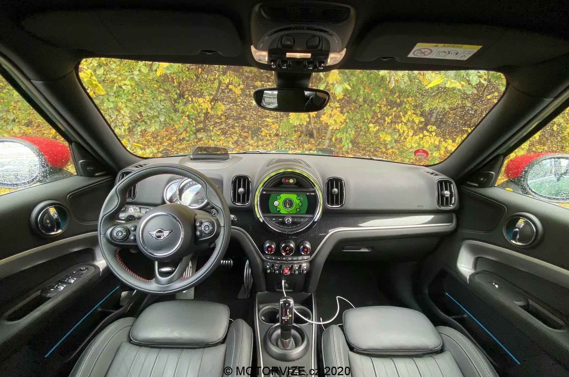 Interior del Mini Countryman 2020 John Cooper Works desde la perspectiva del asiento trasero, centrado en el salpicadero y el área de control del conductor. Se ven el volante con acentos plateados y el logo de la marca, el velocímetro central con pantalla digital, diseño elegante del salpicadero, la palanca de cambios, controles de clima, asientos tapizados en cuero y el espejo retrovisor.