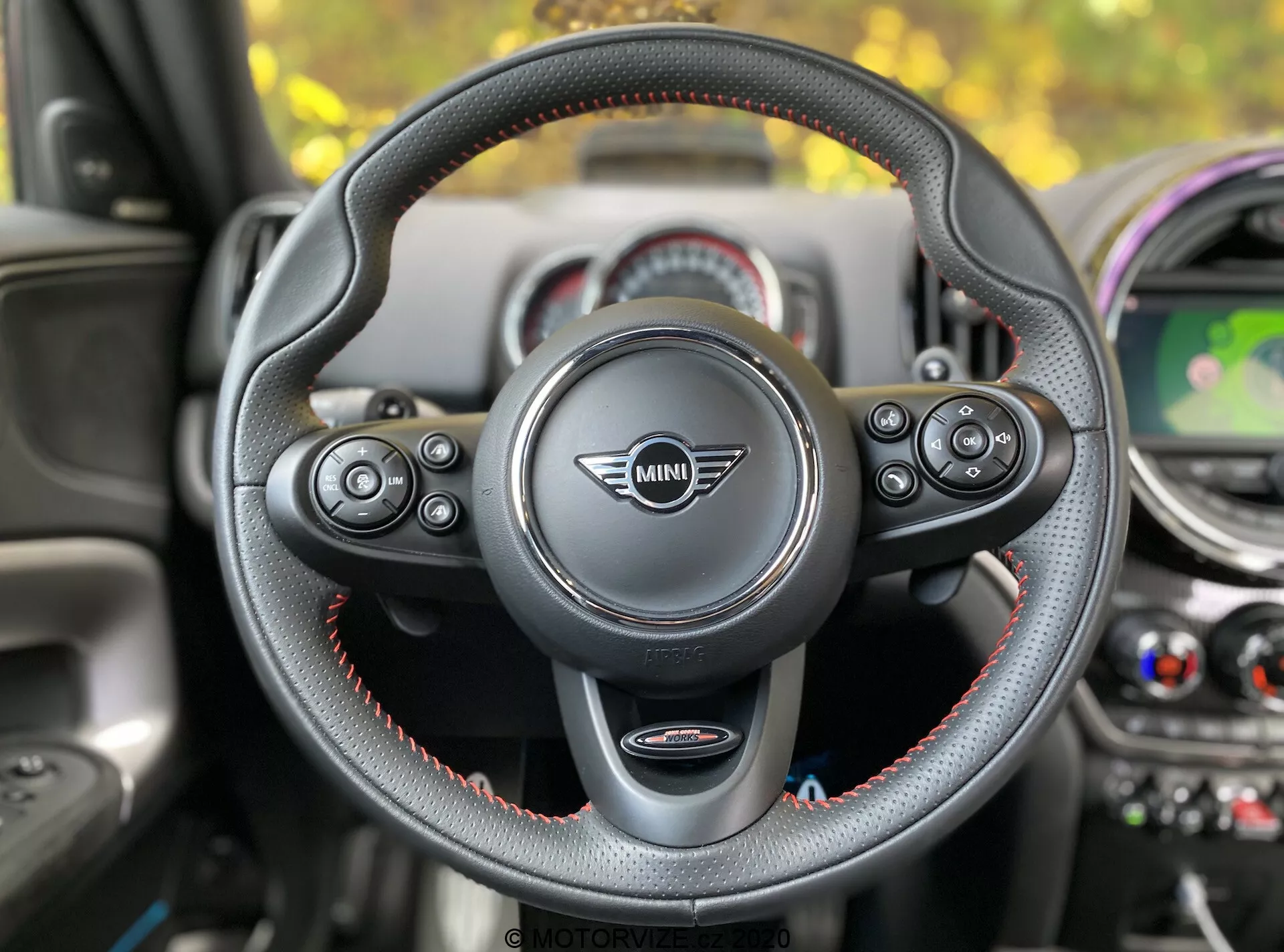 Vista interior de un Mini Countryman John Cooper Works 2020 desde el asiento del conductor, enfocándose en el volante con el logo de Mini en el centro. El volante de tres radios, forrado en cuero con costuras rojas, dispone de controles multifuncionales para audio, teléfono, control por voz y control de crucero. Se ven las levas de cambio, que indican una transmisión automática con selección manual de marchas. En el fondo se incluye el salpicadero con elementos de diseño circular, una pantalla central que muestra un mapa, salidas de aire acondicionado y acentos coloridos.