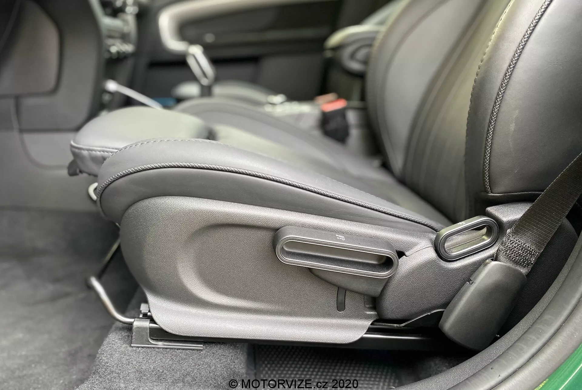 Vista de cerca del interior del Mini Countryman John Cooper Works 2020, destacando la parte inferior lateral del asiento del conductor con mecanismos de ajuste del asiento detallados. El asiento está tapizado en cuero oscuro o cuero sintético con un refuerzo lateral prominente, centrado en la comodidad y la calidad. Se ven los controles manuales de ajuste, incluyendo una manija tipo barra y una perilla redonda, lo que sugiere un diseño ergonómico y ajustabilidad por parte del usuario. También está presente el receptáculo del cinturón de seguridad, indicando atención a la seguridad del pasajero.