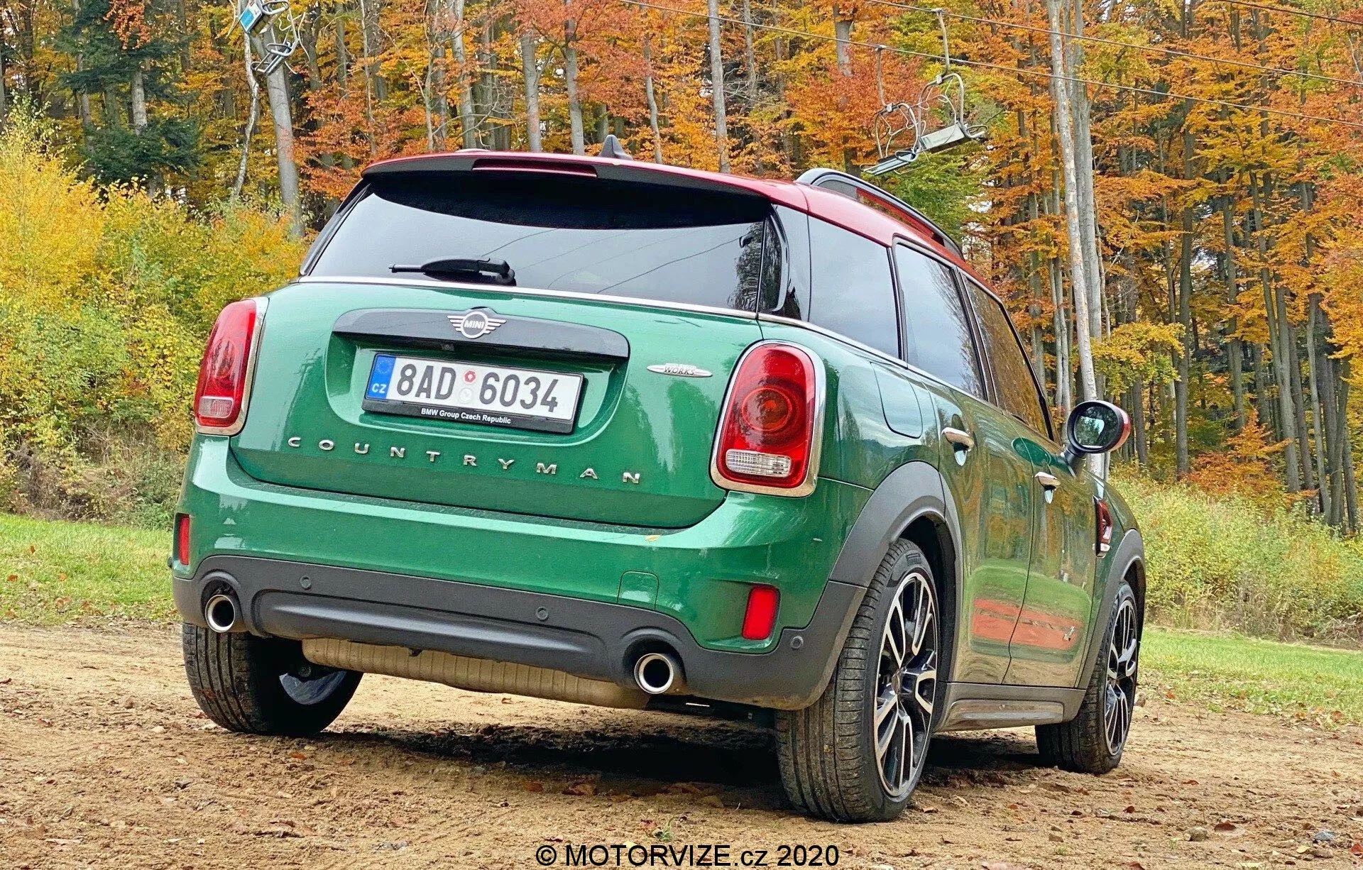 Imagen de un Mini Countryman 2020 modelo John Cooper Works verde estacionado al aire libre en un entorno otoñal, tomada desde un ángulo de tres cuartos traseros. El lado trasero derecho del coche está orientado hacia la cámara y muestra las luces traseras, el parabrisas trasero, la insignia 'COUNTRYMAN', el sistema de escape doble, las ruedas de cinco radios, el techo negro contrastante y los espejos laterales. La imagen también incluye una matrícula trasera, un limpiaparabrisas trasero y una antena para conectividad.