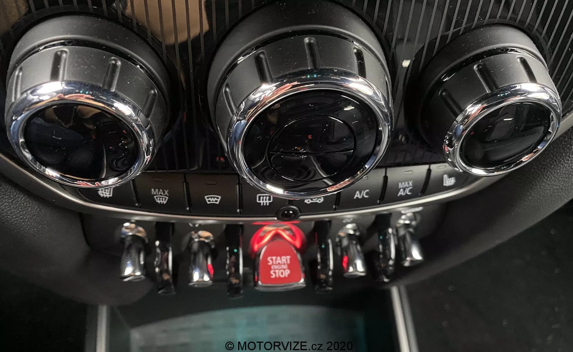 Esta imagen captura una vista de cerca de la consola central en el tablero del Mini Countryman John Cooper Works 2020, destacando el sistema de control de clima y el botón de encendido y apagado del motor. La consola muestra dos ventilaciones de aire circulares con acabados metálicos en la parte superior, un panel de control de clima detallado con tres diales metálicos para temperatura, velocidad del ventilador y dirección del flujo de aire, y varios botones para funciones como descongelado y recirculación de aire. Debajo de los controles hay un botón rojo prominente para el arranque y parada del motor. Las texturas del panel incluyen un diseño deportivo similar a la fibra de carbono alrededor de las ventilaciones de aire y plástico negro estándar para el panel inferior. El ángulo de la foto, desde el frente y ligeramente arriba, resalta el diseño de alta gama y las características avanzadas.