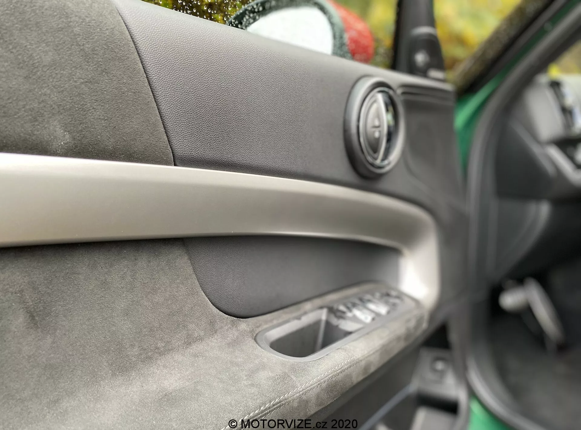 Toma detallada del interior de la puerta de un coche Mini Countryman John Cooper Works 2020. Centrada en altavoz de audio premium con anillo metálico, tira decorativa diagonal, material suave al tacto con costuras visibles, manija interior de la puerta y controles de ventanas eléctricas. Profundidad de campo reducida resaltando la artesanía y la comodidad interior.