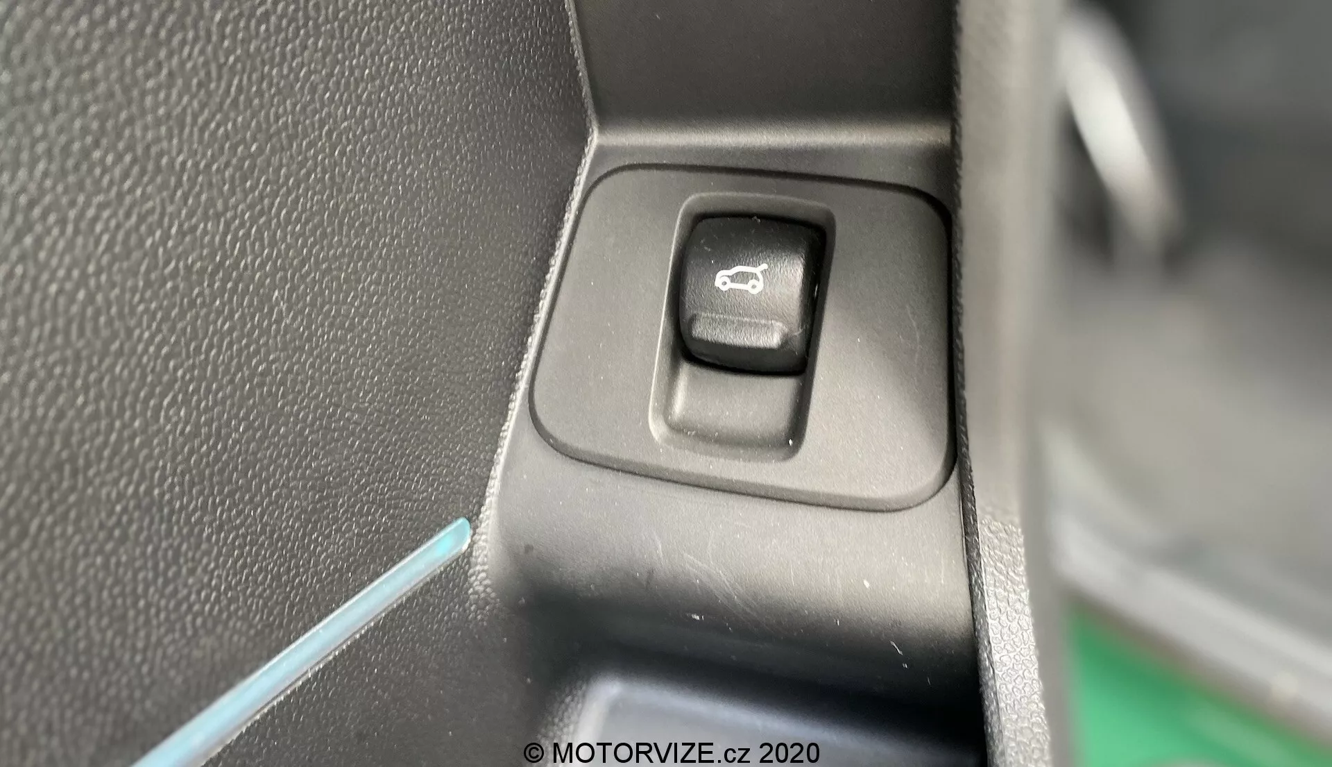 Vista en primer plano del interruptor de control para operar los espejos laterales en el interior del Mini Countryman 2020 John Cooper Works. El interruptor rectangular con esquinas redondeadas, ubicado en un panel plano, está resaltado con un símbolo de espejo. Un lado del interruptor está en sombra, indicando una fuente de luz desde el lado opuesto. El fondo presenta plástico negro texturizado y una delgada línea de luz ambiental azul, contribuyendo a la estética interior del vehículo.
