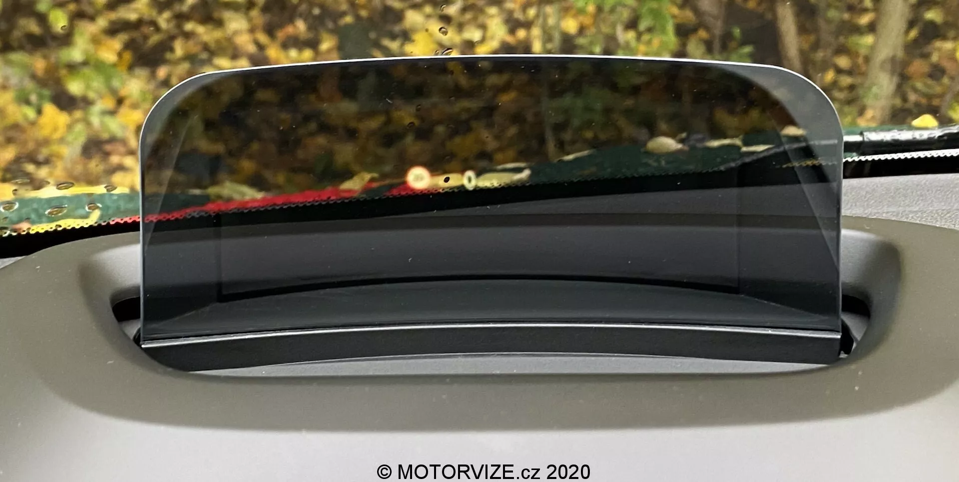 Imagen mostrando el head-up display (HUD) del Mini Countryman 2020, proyectado sobre una pantalla transparente encima del tablero desde la perspectiva del conductor. El panel claro y semejante al vidrio del HUD se integra perfectamente con el diseño interior, siendo redondeado y ligeramente inclinado para una visibilidad óptima. El fondo fuera del parabrisas está borroso, enfatizando el foco del HUD en presentar información crucial para la conducción.