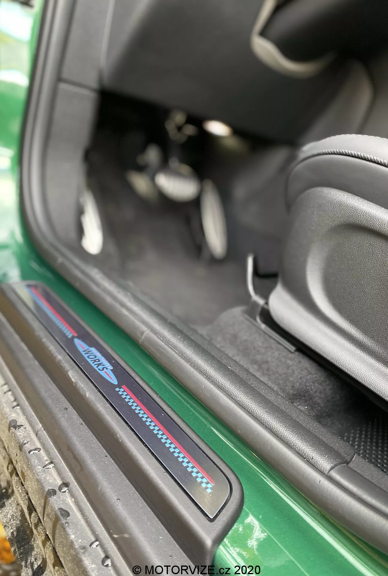 Vista de primer plano del área del umbral de la puerta del Mini Countryman John Cooper Works 2020 desde un ángulo oblicuo enfocado en el umbral del lado del pasajero. La imagen, capturada desde fuera del vehículo, destaca la parte inferior del interior del coche, incluyendo la placa del umbral con acentos en azul y rojo, alfombras negras, el costado deportivo del asiento negro, una vista parcial de los pedales y un área de umbral bien cuidada. Los elementos exteriores visibles incluyen una falda lateral verde y la carrocería, realzando el atractivo estético. La marca de agua 'MOTORVIZE.cz 2020' sugiere que la foto se tomó en 2020 para una reseña automotriz.