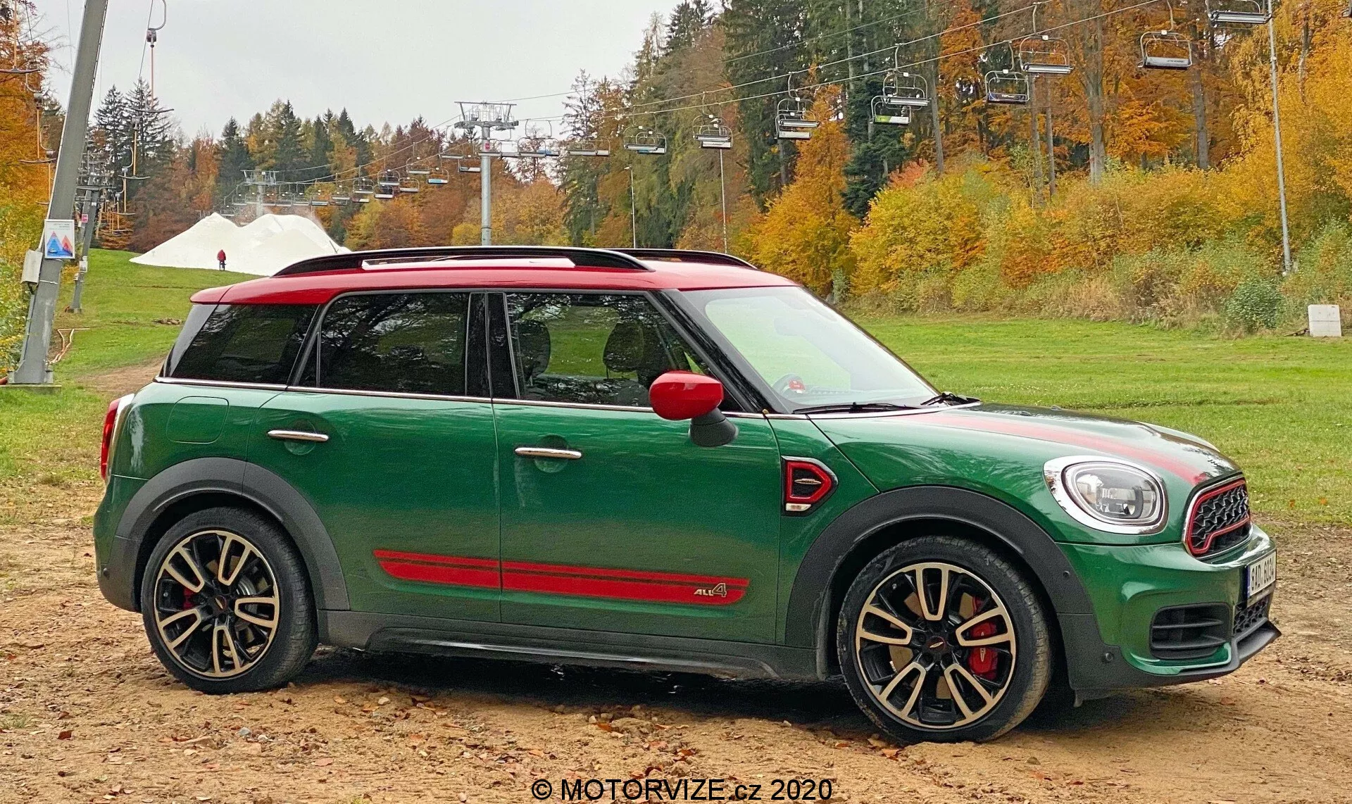 Un Mini Countryman John Cooper Works 2020 estacionado sobre suelo húmedo con una carrocería verde y techo rojo. Vista de perfil lateral bajo cielos nublados. Características incluyen espejos laterales rojos, ruedas de aleación deportivas, una distintiva raya roja en la parte baja de las puertas, un indicador lateral delante del arco de la rueda delantera y una antena corta en el techo. El fondo muestra una zona arbolada con colores otoñales y una estructura blanca distante.
