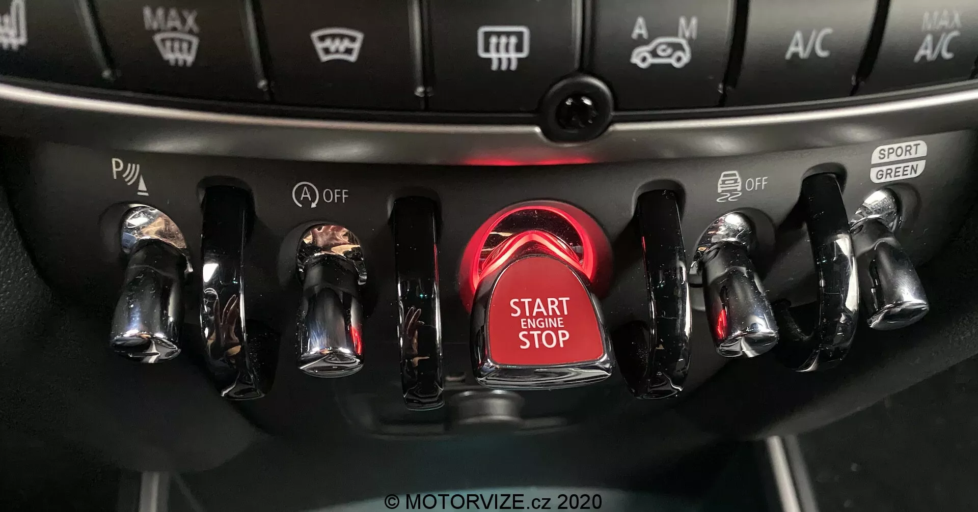 Vista de cerca de la consola central del Mini Countryman John Cooper Works 2020, enfocándose en el área de encendido con un botón de arranque/parada de motor destacado y rodeado por un anillo rojo brillante, con interruptores metálicos acabados en cromo a ambos lados para varios controles del vehículo, incluyendo modos de conducción y configuraciones de clima.