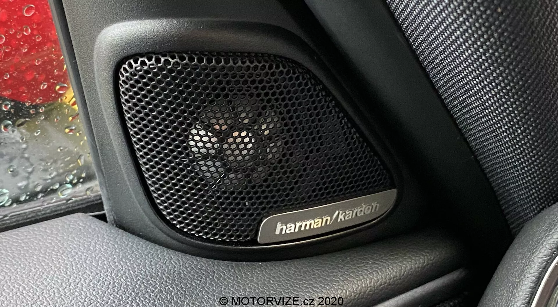 Vista en primer plano del sistema de altavoces Harman Kardon en el interior de la puerta de un Mini Countryman 2020 John Cooper Works. La fotografía se centra en la rejilla perforada del altavoz, diseñada para la transparencia acústica y la protección, situada contra un interior con textura similar al cuero. Se ven gotas de lluvia en la ventana del coche, añadiendo contexto a la imagen. El sistema de audio premium señaliza características tecnológicas clave, mostrando el lujo y durabilidad del diseño del vehículo.