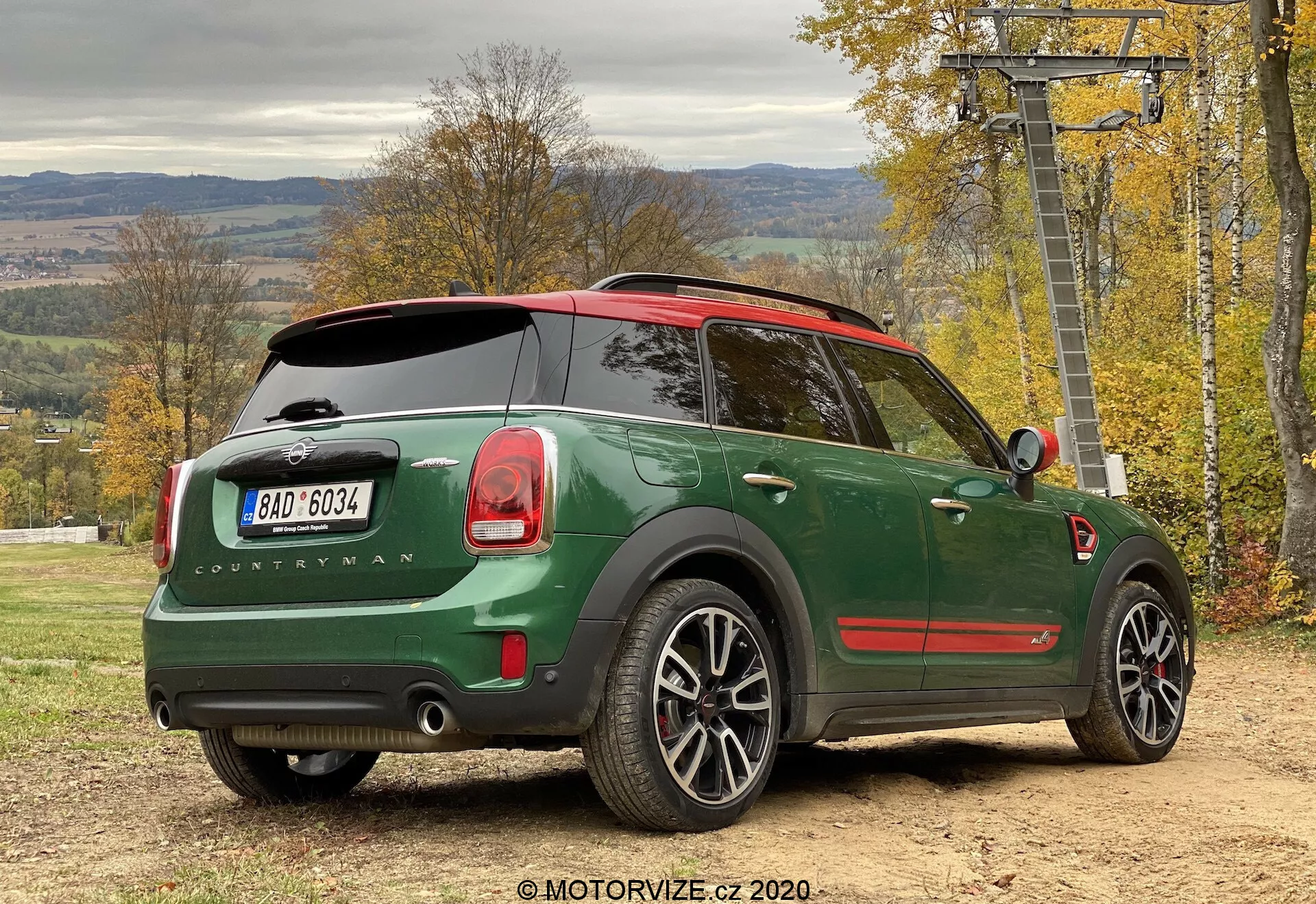Vista trasera de tres cuartos de un Mini Cooper Countryman John Cooper Works 2020 verde estacionado al aire libre, destacando el diseño del coche desde la perspectiva lateral trasera. Los detalles notables incluyen una carrocería verde oscuro con techo rojo, insignia 'Countryman', logotipo de Mini en el portón trasero, luces traseras distintivas, sistema de escape doble, llantas de aleación, techo panorámico, limpiaparabrisas trasero, ventanas traseras tintadas, sensores de aparcamiento y un espejo lateral con luz direccional integrada. El vehículo está en una superficie sin pavimentar con follaje otoñal y una estructura de telesilla en el fondo, lo que sugiere un estilo de vida activo y al aire libre.