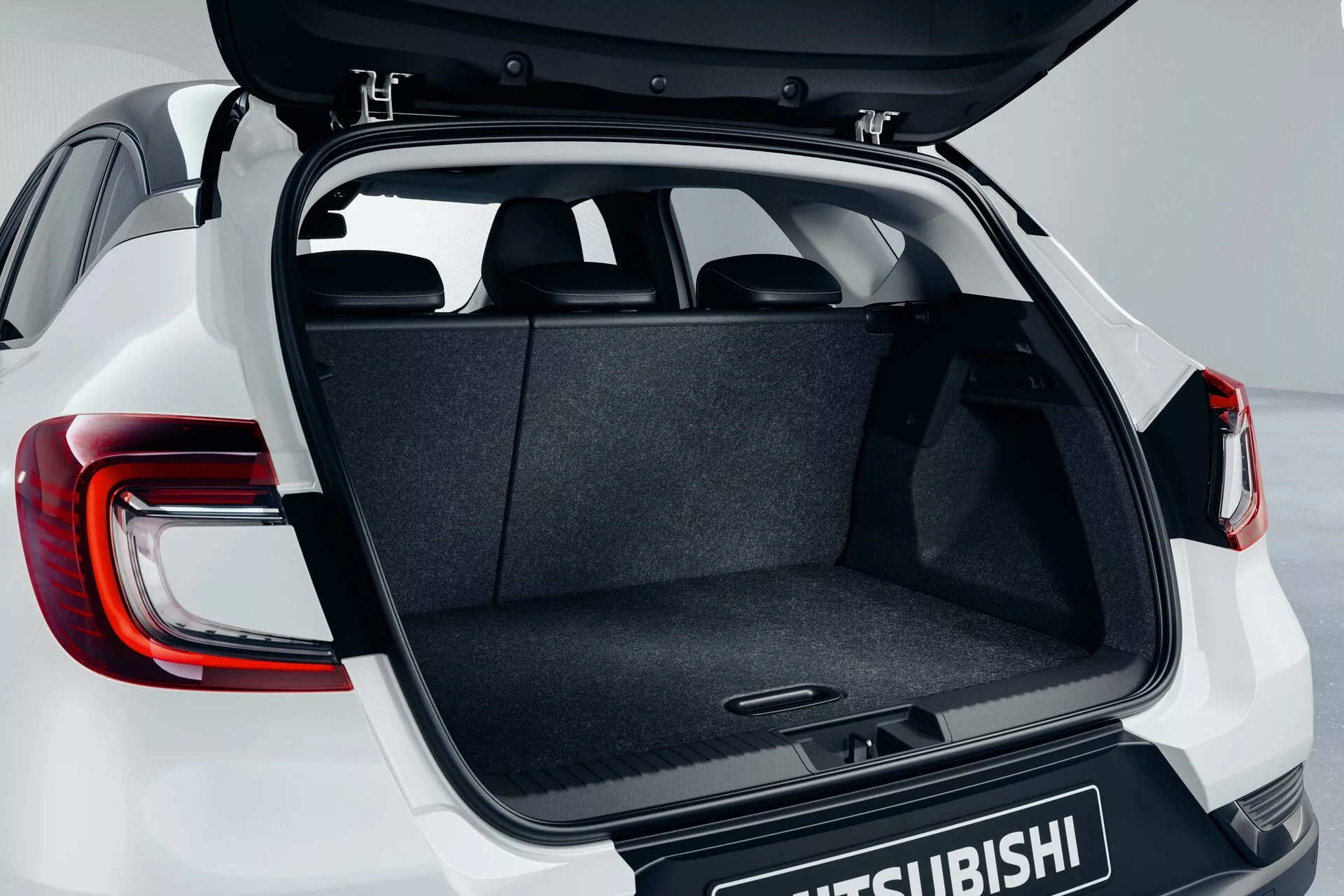 Maletero del Mitsubishi ASX 2023 (2ª generación) mostrando su compuerta abierta desde un ángulo ligeramente elevado, destacando el espacio de carga alfombrado, el mecanismo de bisagra, el piso plano y las luces traseras LED modernas parcialmente visibles con el logotipo de Mitsubishi en la puerta del maletero.