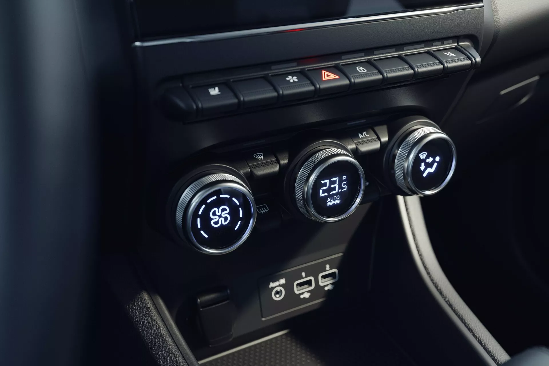 Vista cercana del sistema de control de clima interior del Mitsubishi ASX 2023 (2ª generación). La imagen, tomada desde un ángulo elevado, se enfoca en la consola central con tres diales de control de temperatura: el izquierdo en 'LO' y el derecho en '23.5'. El dial central parece controlar la velocidad o dirección del ventilador. Arriba hay botones para desempañar, luces de emergencia y circulación de aire. Abajo, hay puertos USB modernos. El diseño es minimalista con superficies negras y elegantes.