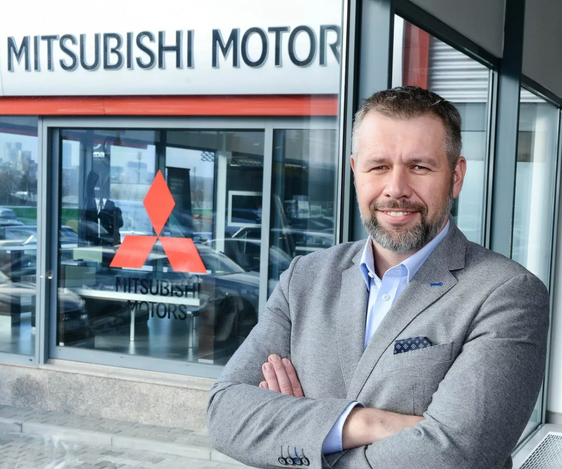 Persona de pie con los brazos cruzados frente a un concesionario de Mitsubishi Motors, vehículo parcialmente visible a través de la ventana de cristal sugiriendo un modelo de exposición en el fondo