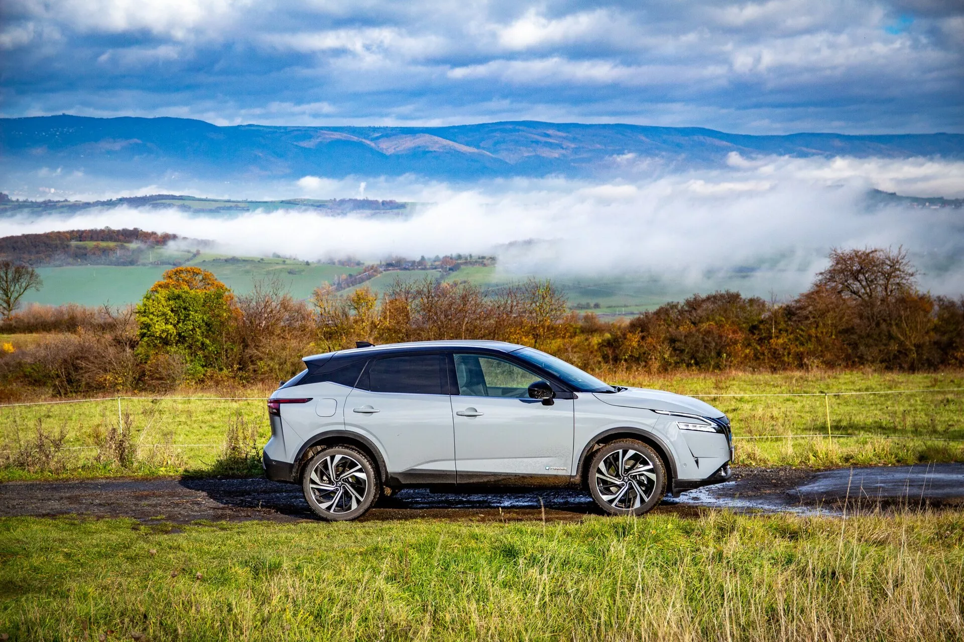 Vista de perfil lateral de un Nissan Qashqai 2021, un SUV moderno con un acabado brillante de color plata o gris claro, ruedas de aleación de múltiples radios, ventanas tintadas y una línea de techo inclinada, estacionado en un camino rural mojado con charcos. El diseño elegante del coche, las unidades de iluminación avanzadas y las señales de giro integradas en los espejos laterales son claramente visibles contra un fondo de colinas ondulantes y nubes bajas.