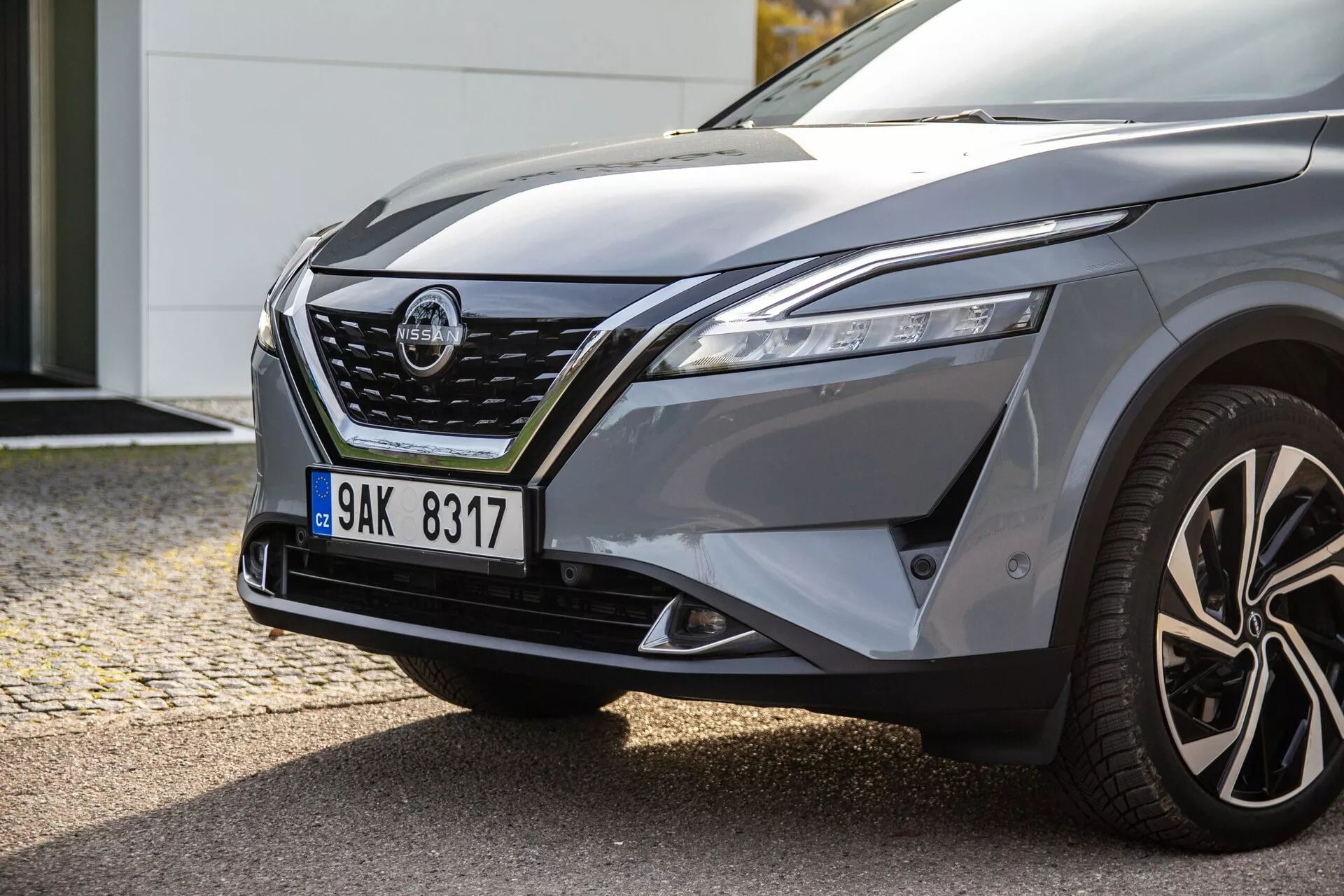 Primer plano del lado delantero izquierdo de un coche moderno, que muestra faros LED elegantes, una parrilla prominente con acentos cromados, un capó esculpido, un guardabarros abultado, llantas de aleación de múltiples radios y un espejo lateral del conductor con señales de giro integradas, reflejando el diseño dinámico y las potenciales capacidades de manejo del coche.