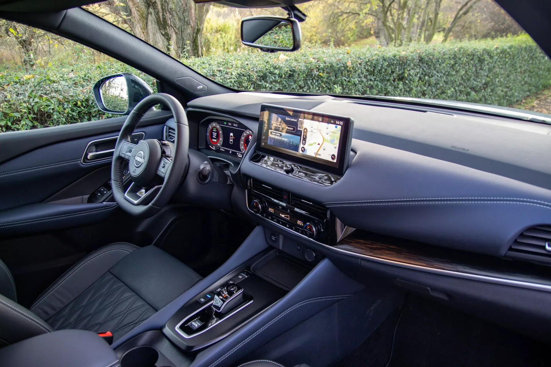 Vista interior del Nissan Qashqai 2021 desde la perspectiva del asiento trasero, mostrando la moderna cabina delantera con volante multifuncional, panel de instrumentos totalmente digital y una gran pantalla táctil de infoentretenimiento. La consola central presenta una palanca de cambios automática, botones de control de clima y opciones de conectividad, con asientos delanteros tapizados en cuero y materiales premium en el tablero y la consola.