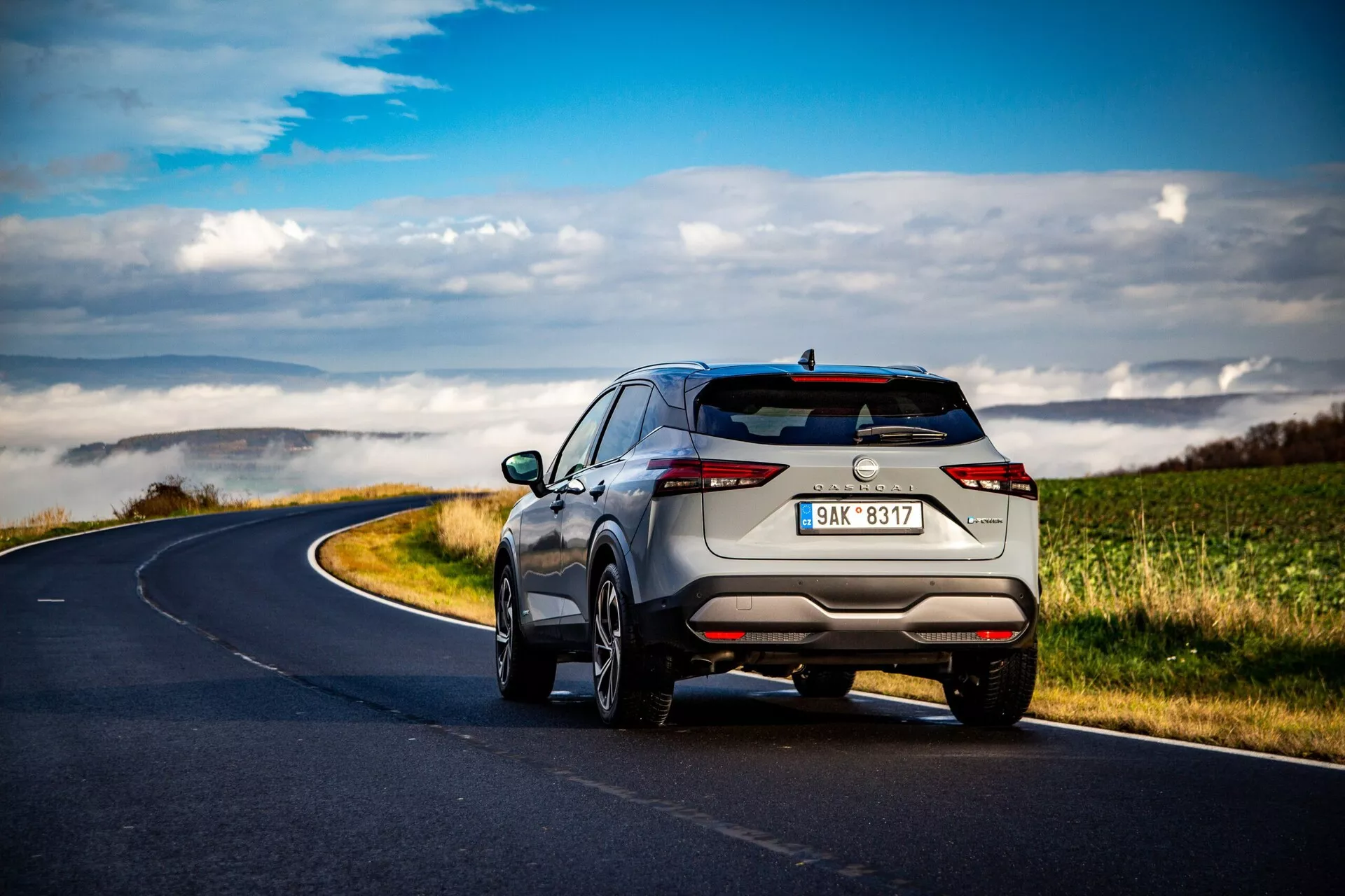 SUV Nissan Qashqai 2021 capturado desde un ángulo trasero de tres cuartos en un camino rural serpenteante bajo nubes dispersas, mostrando un diseño elegante y moderno con luces traseras delgadas horizontales, tubos de escape discretos, llantas de aleación de múltiples radios, rieles en el techo y sensores de estacionamiento avanzados. El color metálico claro resalta su sensación premium.