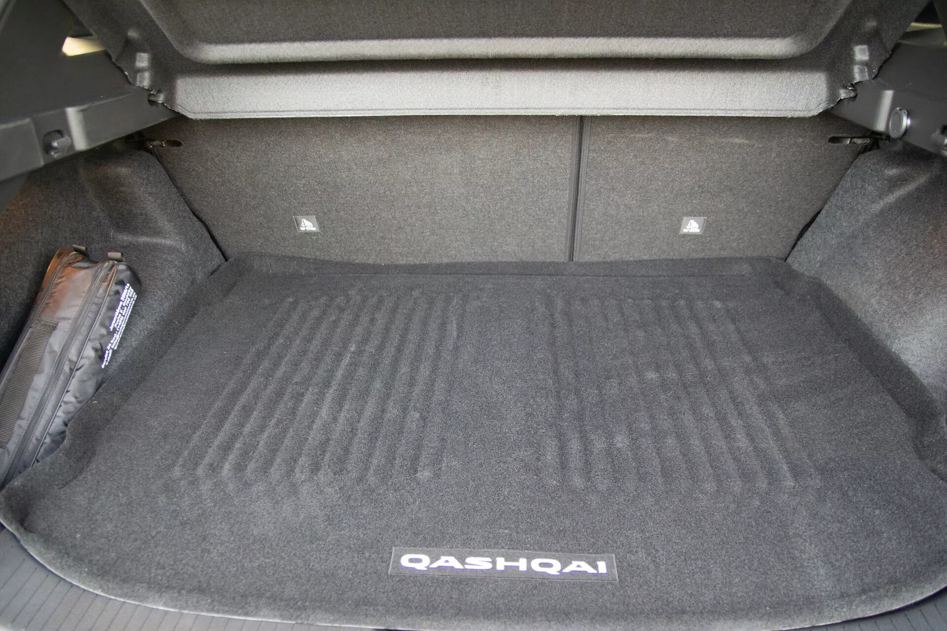Imagen que muestra el espacio del maletero abierto de un Nissan Qashqai 2021 desde un ángulo trasero. El maletero, vacío de carga, revela su superficie alfombrada, paredes laterales y los asientos traseros en posición vertical. Un área ligeramente elevada en la parte delantera del maletero sugiere componentes mecánicos subyacentes o soporte para los asientos traseros. En la parte trasera del maletero se puede ver una partición de almacenamiento menor. A la izquierda, hay un artículo de equipo de emergencia, posiblemente un gato para neumáticos o un botiquín de primeros auxilios. La imagen destaca la capacidad espaciosa y el diseño funcional del maletero del vehículo.